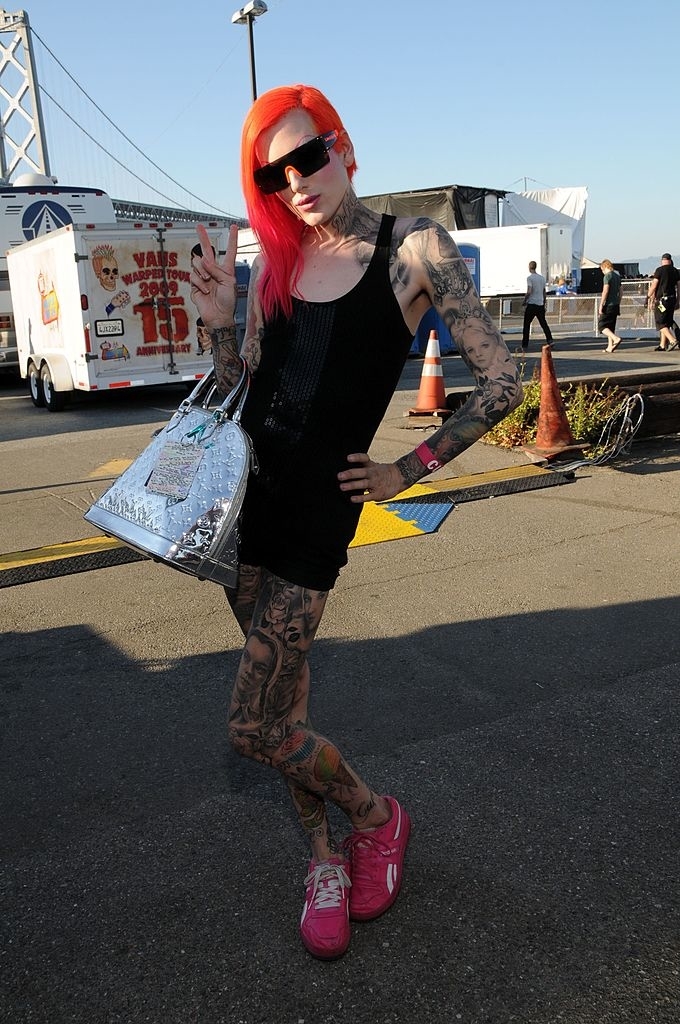 Person in a black sleeveless outfit with visible tattoos, bright red hair, sunglasses, holding a silver bag, and making a peace sign gesture
