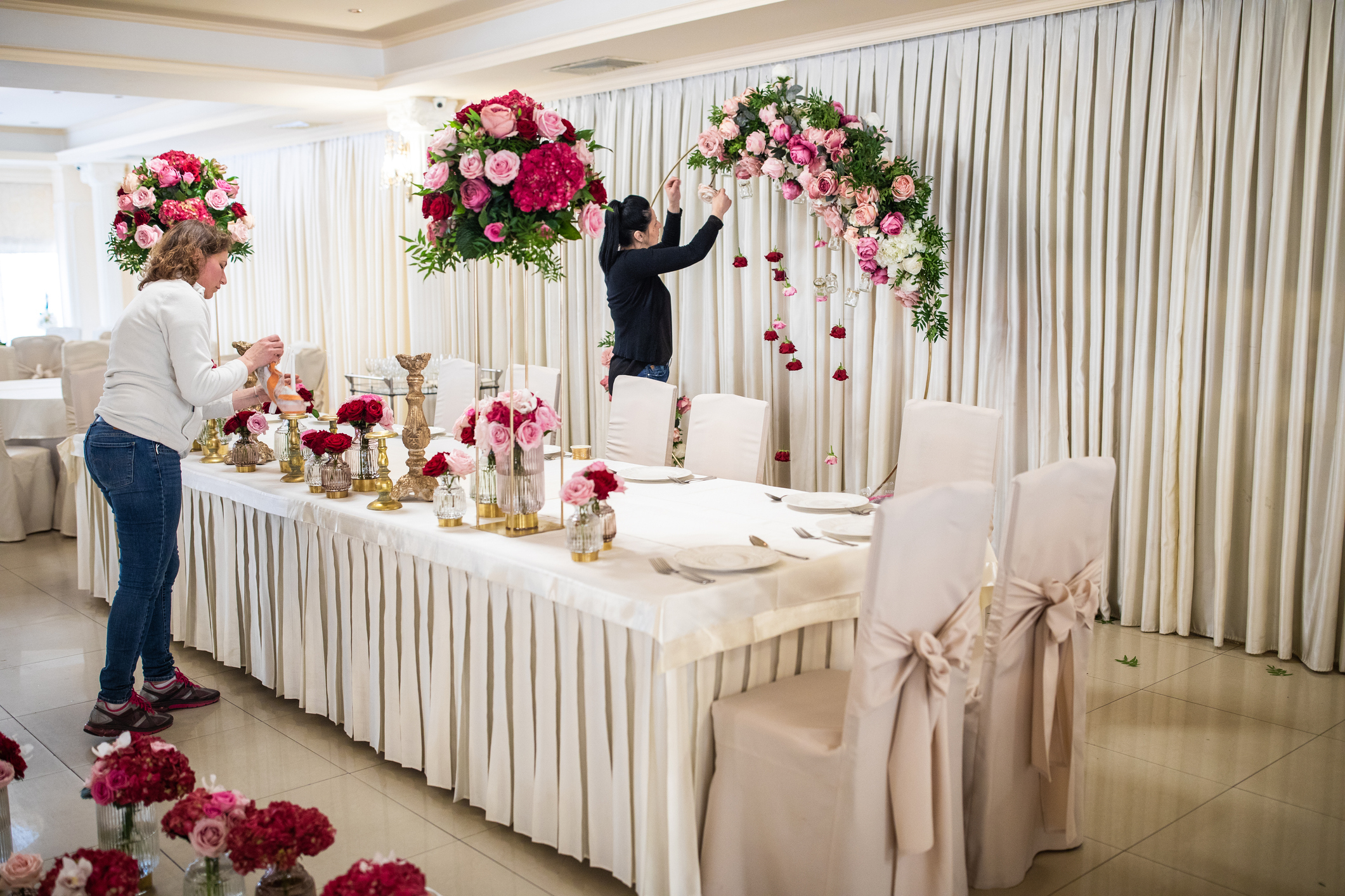 Two people are decorating a banquet hall with flower arrangements for a wedding