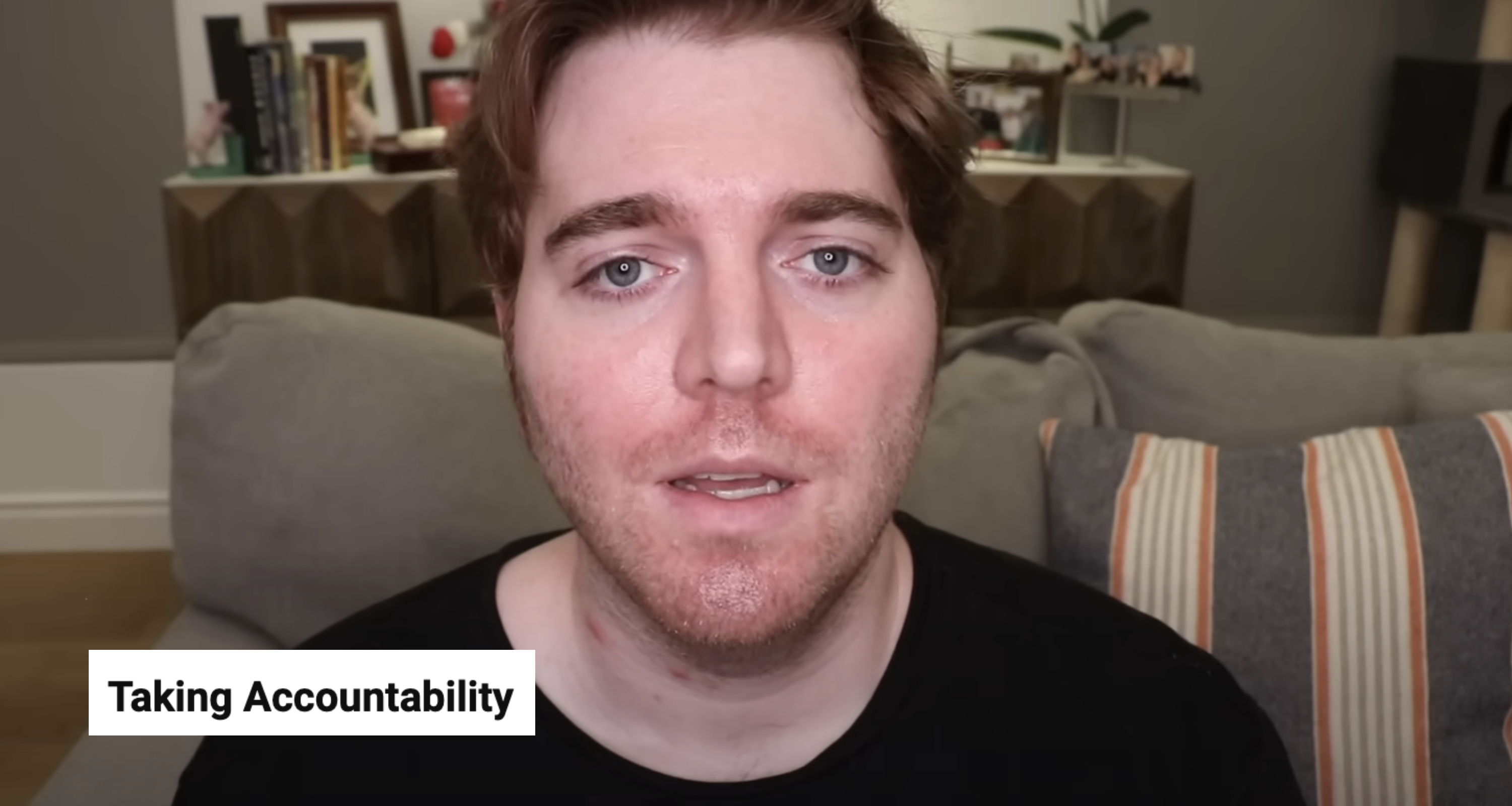 A person with short hair, wearing a casual black shirt, appears in an indoor setting with a couch and bookshelf in the background. Faces slightly concerned