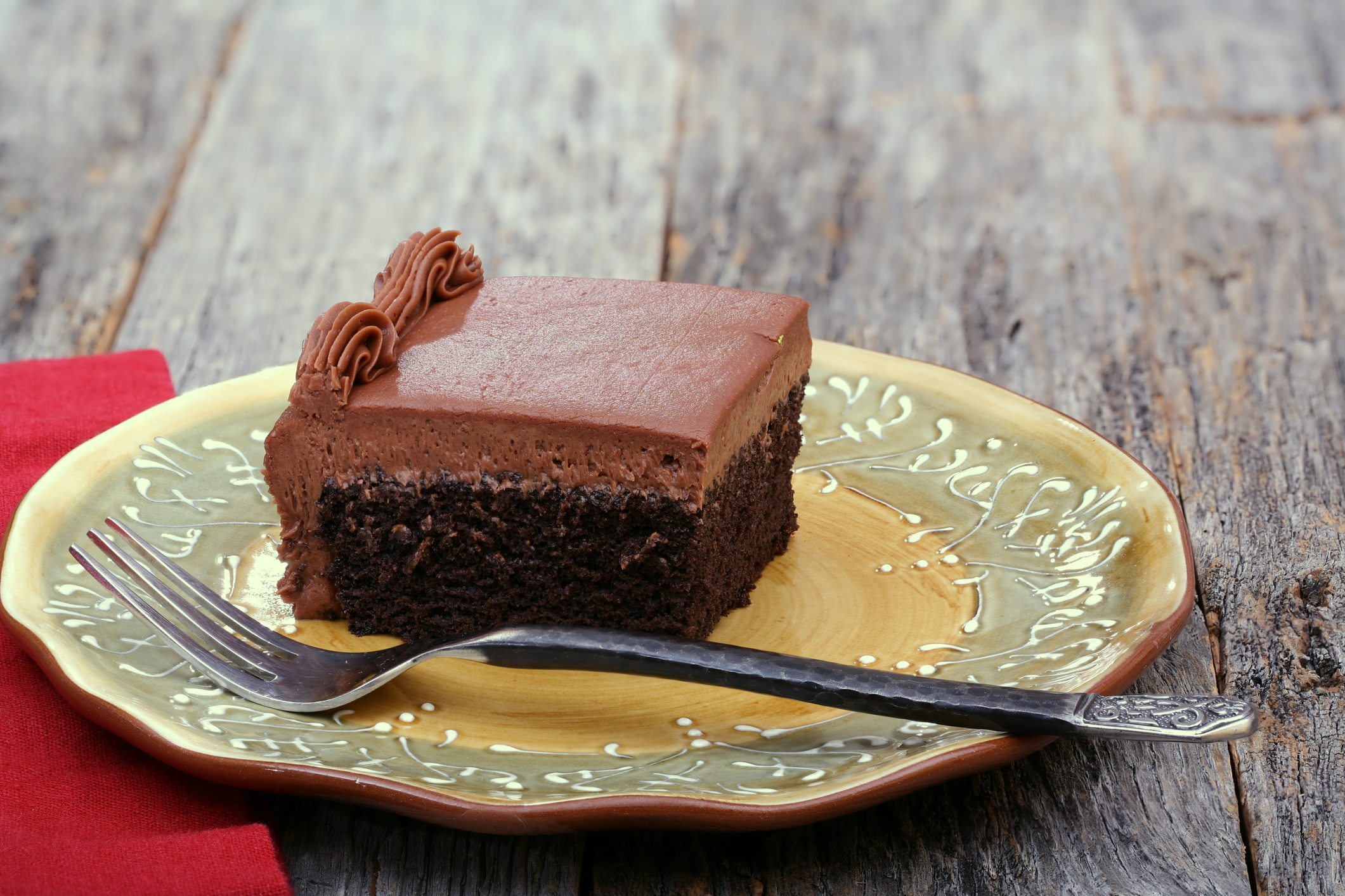 A slice of chocolate cake with icing on a decorative plate, accompanied by a fork on the side