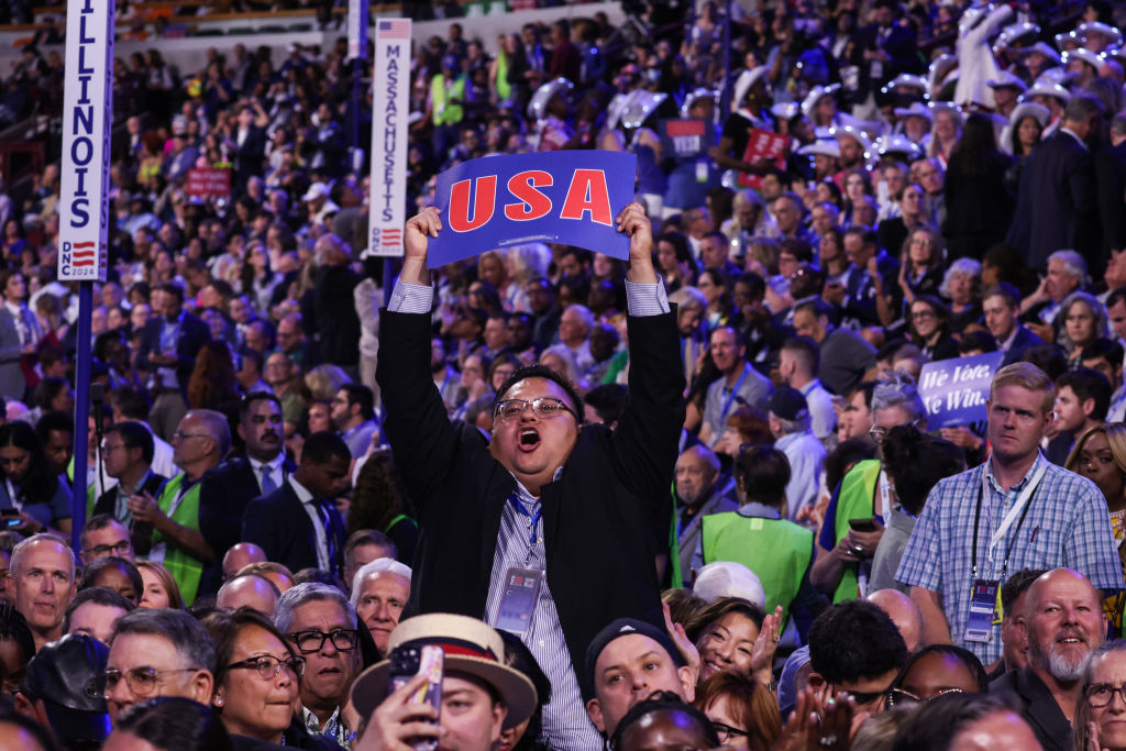 A man excitedly holds a &quot;USA&quot; sign in a crowd at a political event. People around him display various signs and expressions
