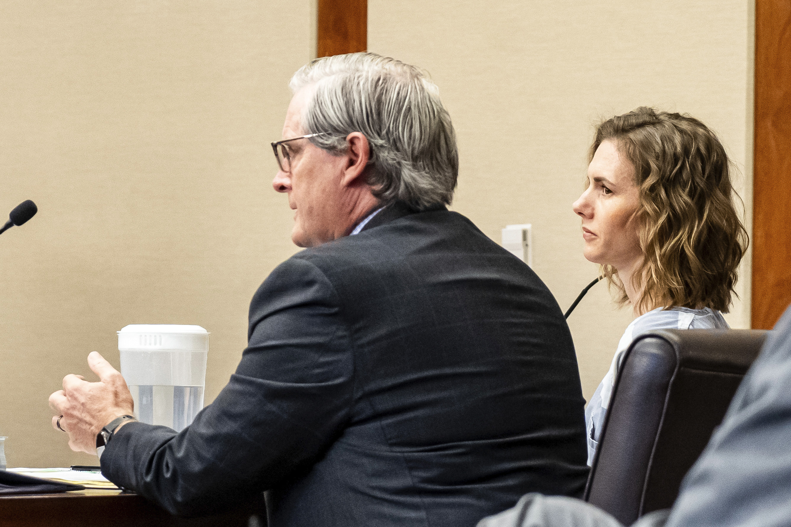 Two people sit at a table in a formal setting, speaking into microphones