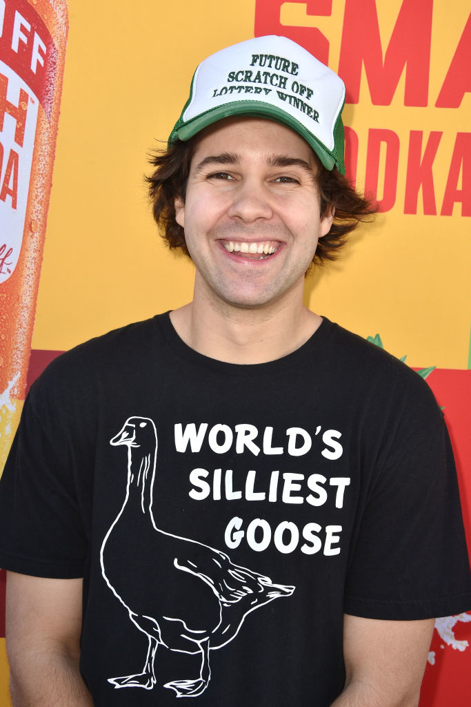 David Dobrik wearing a t-shirt with "World's Silliest Goose" print and a hat that reads "Future Scratch Off Lottery Winner," smiles at an event