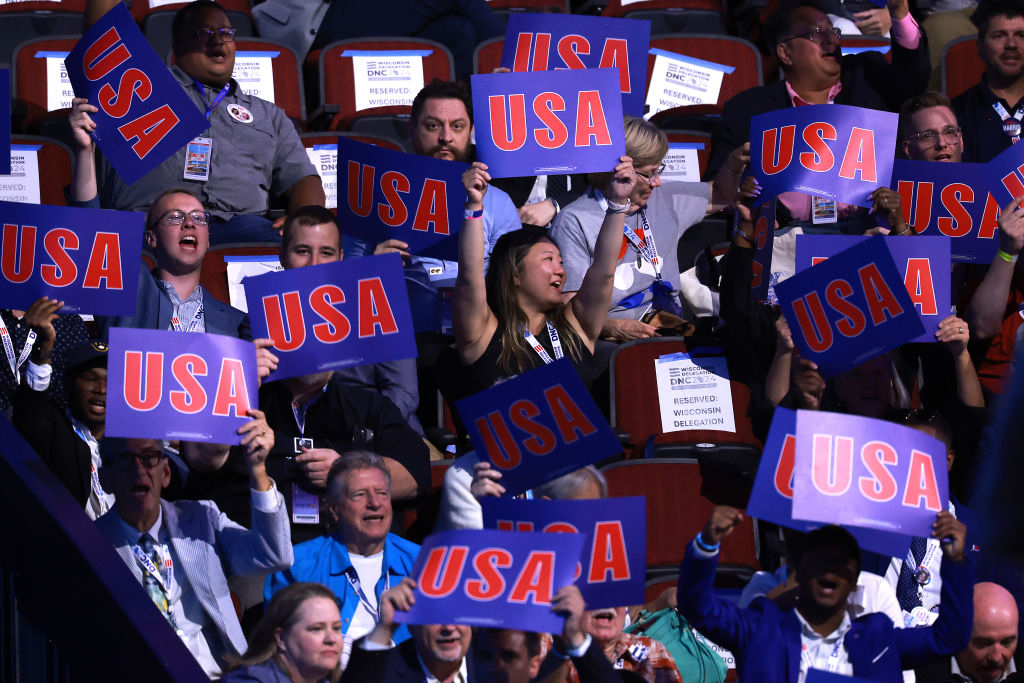 A cheering crowd holds &quot;USA&quot; signs at an event, with diverse attendees enthusiastically showing support. Names of persons unknown