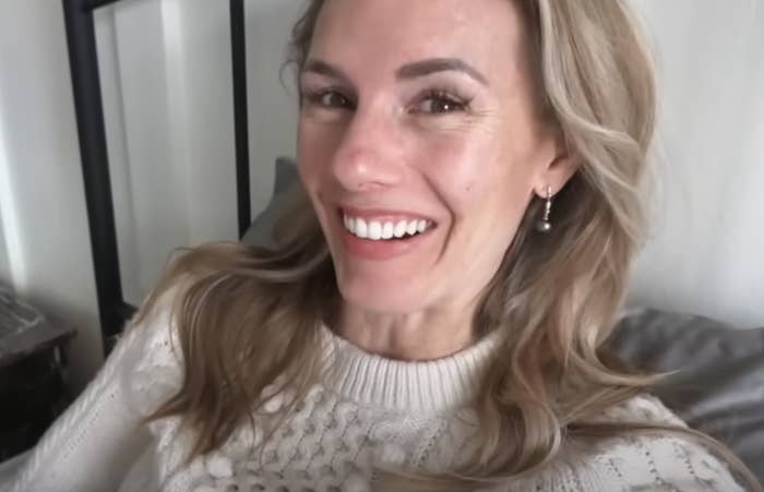 A smiling woman with wavy hair and a knitted sweater leans back on a cushion