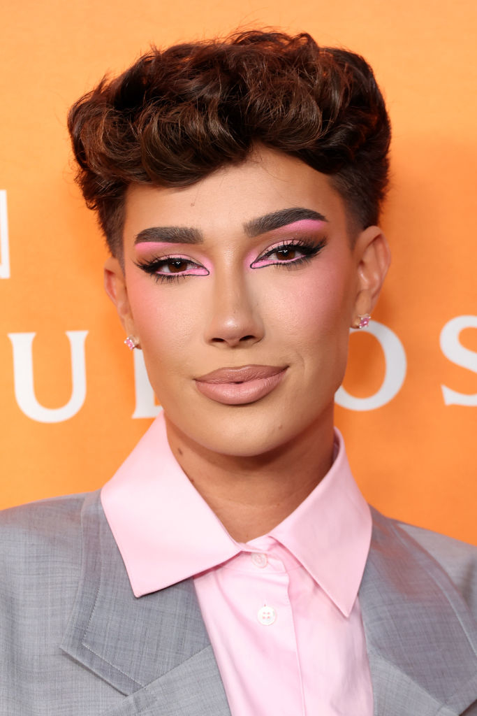 James Charles at an event, wearing a light pink button-up shirt under a light grey jacket, with bold makeup featuring pink eyeshadow and winged eyeliner