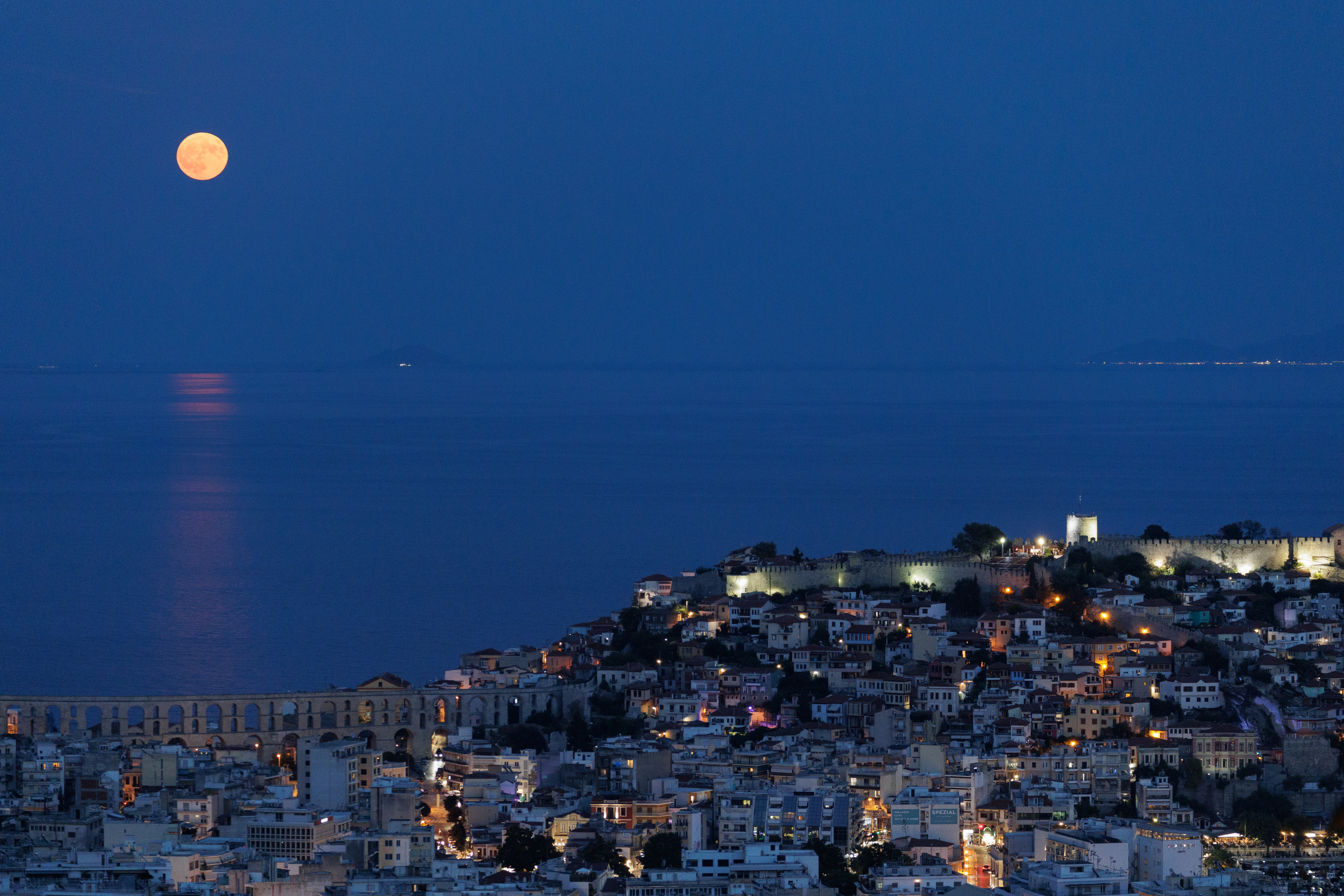 A full moon rises over a coastal city, illuminating the sea and urban landscape below. The image captures the serene nighttime cityscape