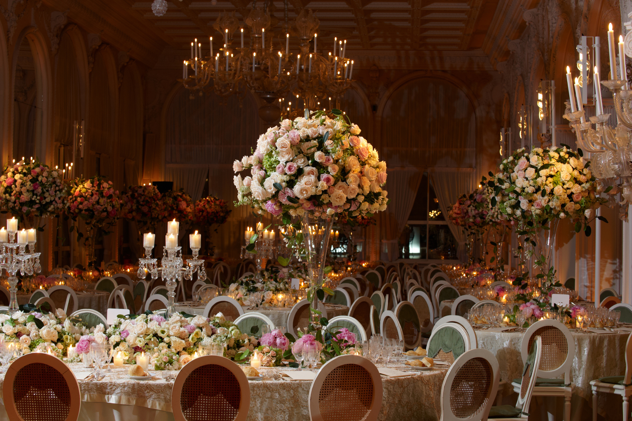 Elegant ballroom with chandeliers and floral arrangements set for a formal event, featuring decorative dining tables and ornate candle holders