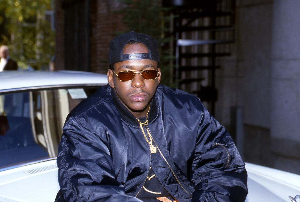 Bobby Brown in a dark jacket and cap, leaning against a car, wearing sunglasses and gold jewelry