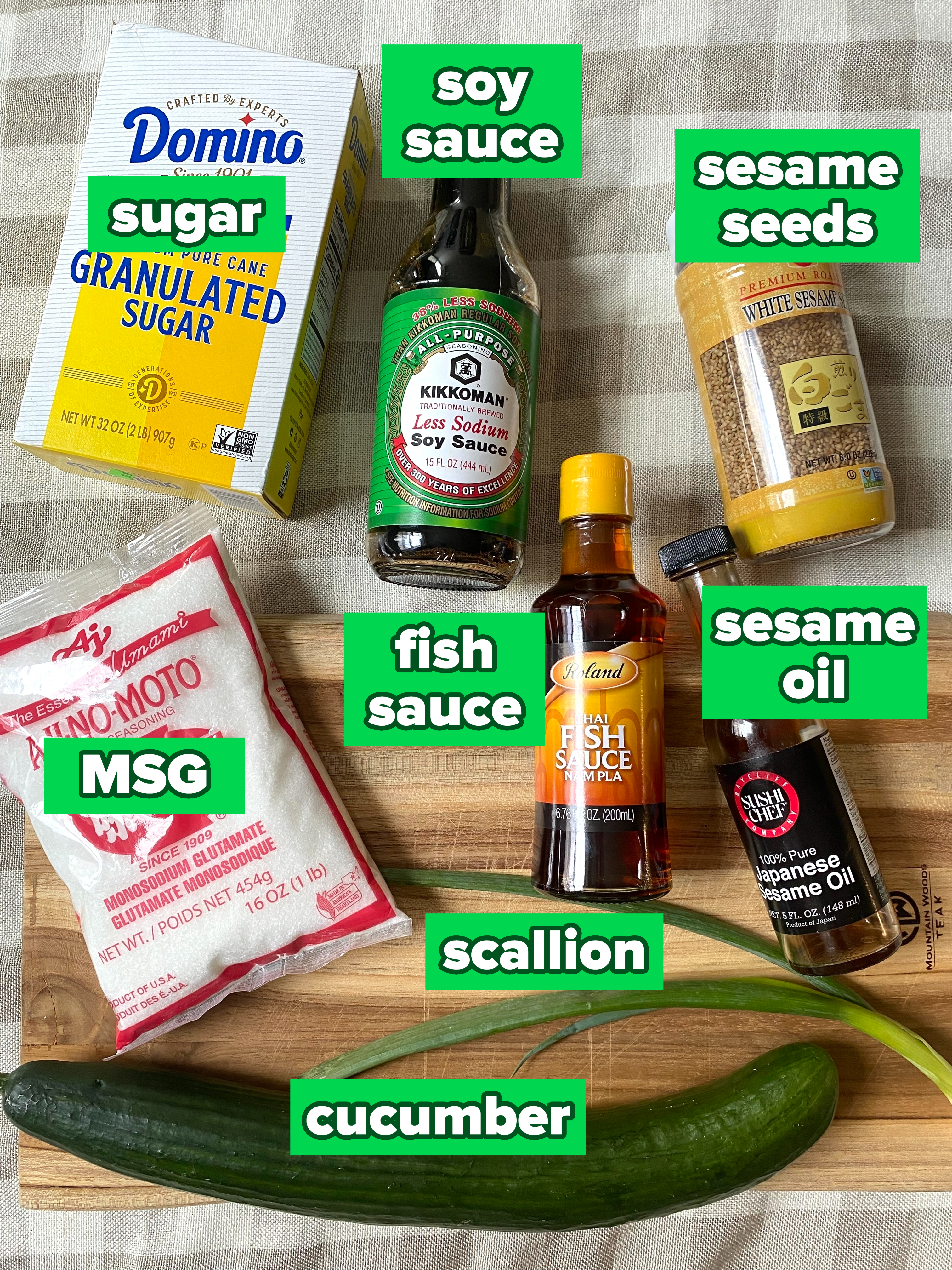Various cooking ingredients including granulated sugar, soy sauce, sesame seeds, fish sauce, MSG, sesame oil, a cucumber, and green onion on a wood board