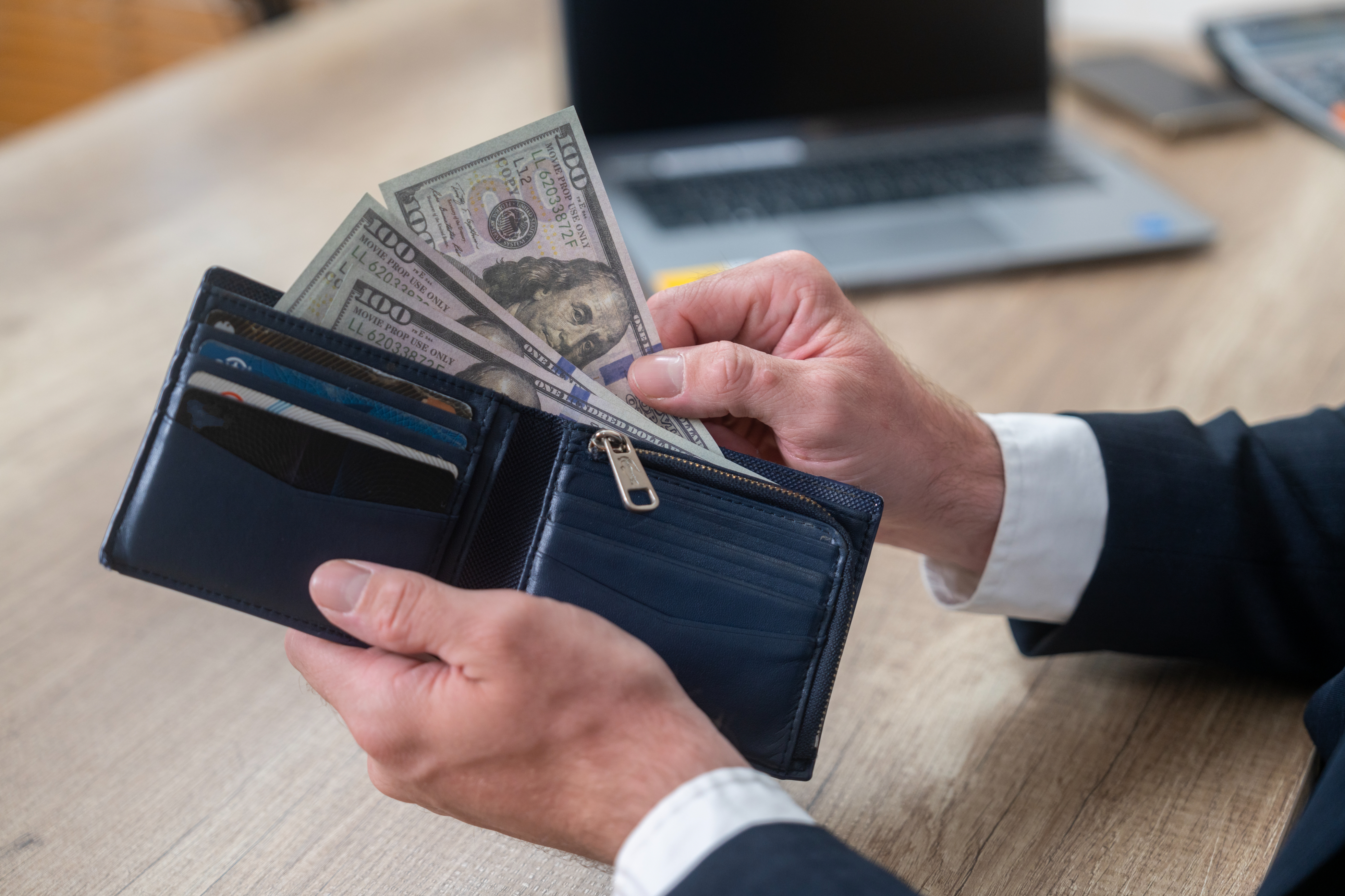 Person holding a wallet containing several credit cards and counting U.S. dollar bills, sitting at a desk with a laptop in the background