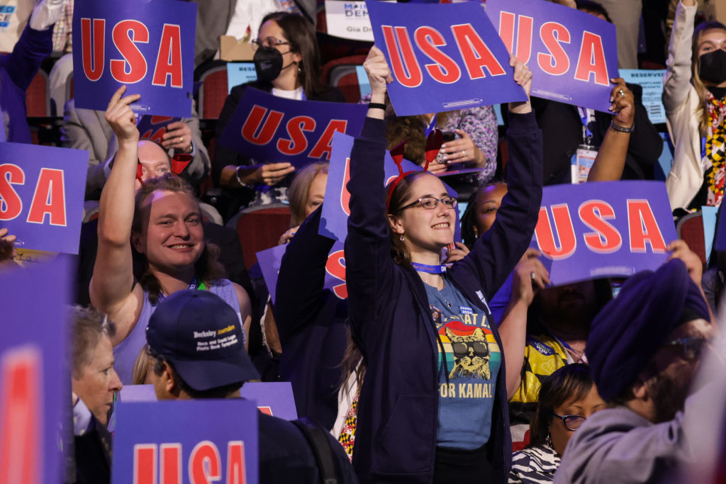 A crowd of people at an event hold up signs reading &quot;USA.&quot; Multiple people are smiling and cheering as they lift their signs high