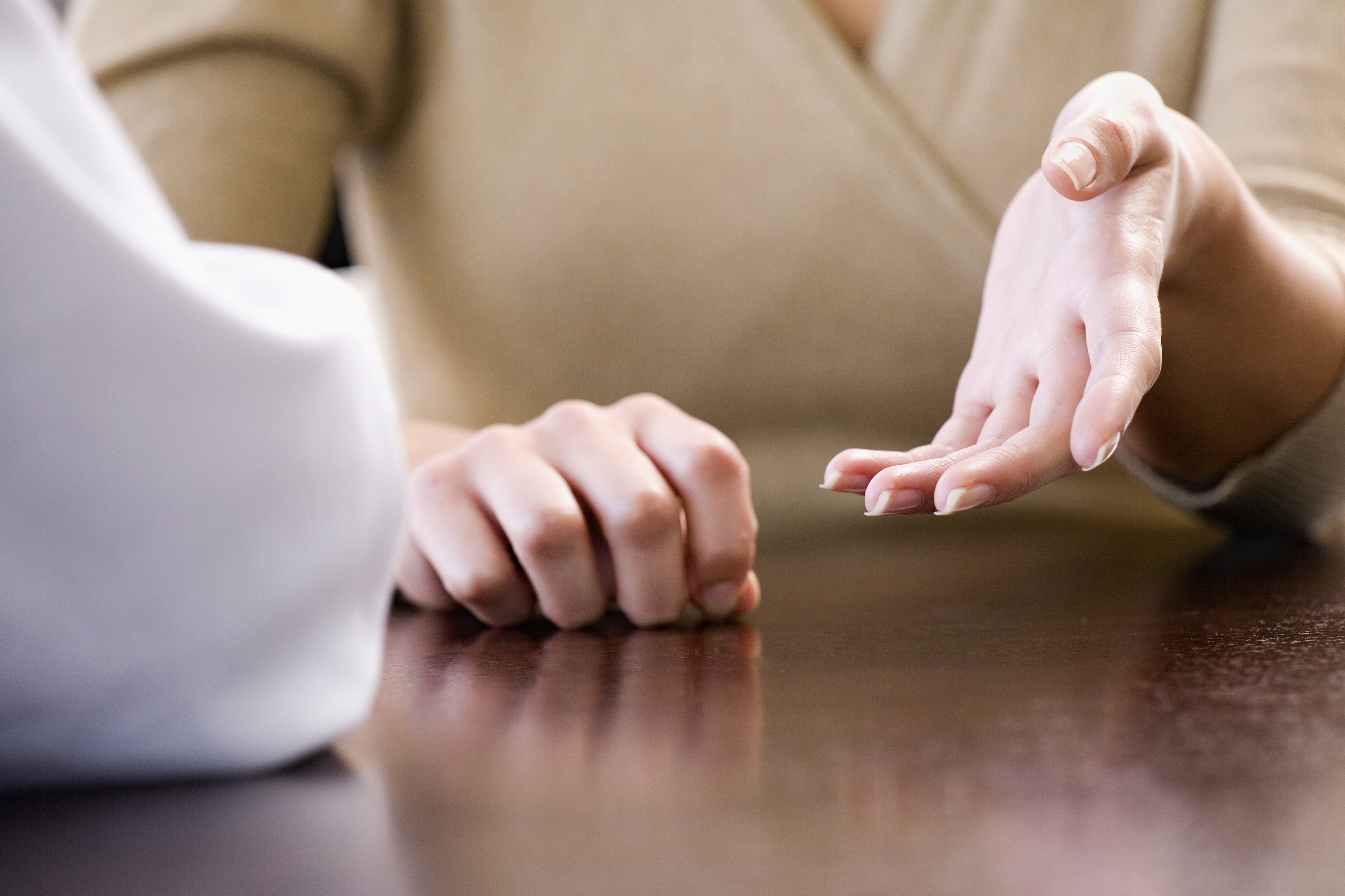 A person sits at a table with another, their hands gesturing as if in conversation