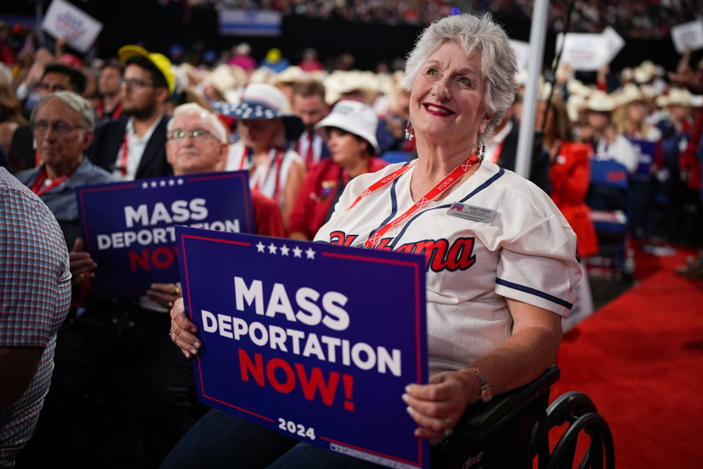 A woman in a crowd at an event holding a sign that reads &quot;Mass Deportation Now!&quot; The crowd includes people in casual attire and hats