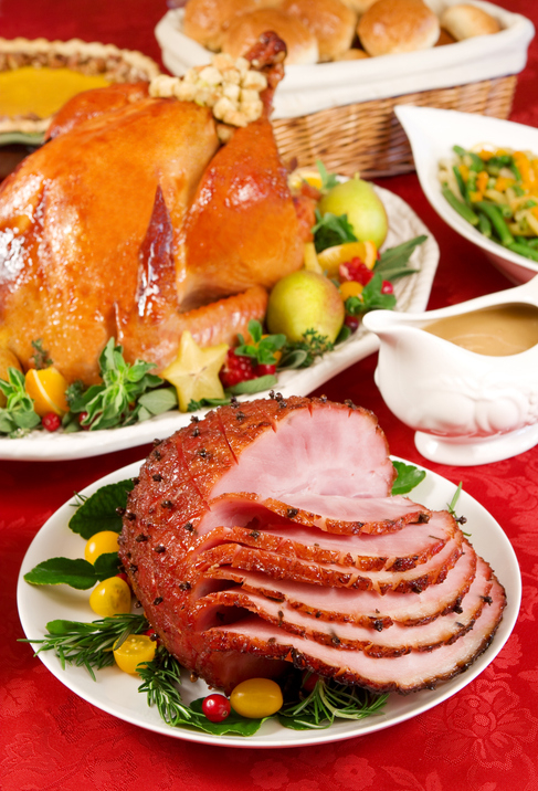 A festive dinner setting with a glazed ham centerpiece, a roast turkey, dinner rolls in a basket, green beans, and a gravy boat on a red tablecloth