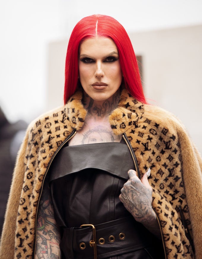 Jeffree Star, with bright red hair, wears a stylish black leather outfit and a patterned fur coat on a red carpet, showcasing tattoos