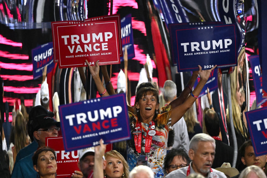 A crowd holds signs supporting Trump and Vance at a political rally. One woman stands out, enthusiastically raising a &quot;Trump&quot; sign above her head