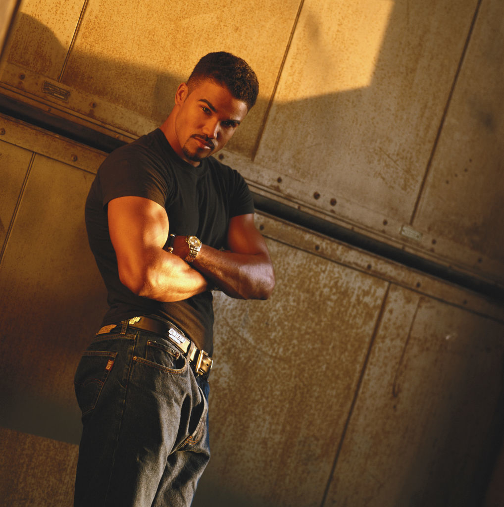 A man stands against a metal wall, arms crossed, wearing a fitted black t-shirt, jeans, and a belt