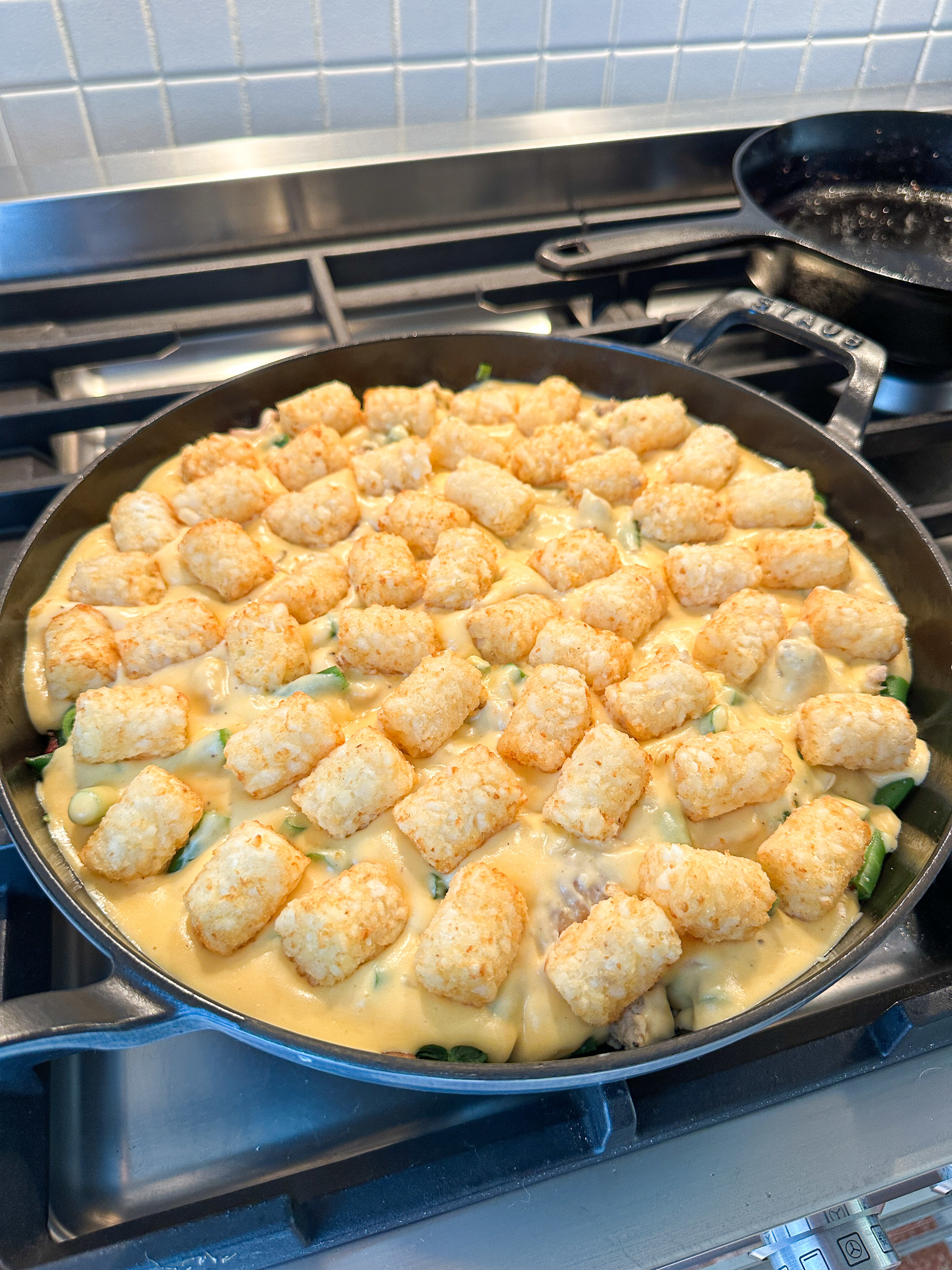 A skillet on a stove filled with a creamy casserole topped with tater tots, in the process of cooking