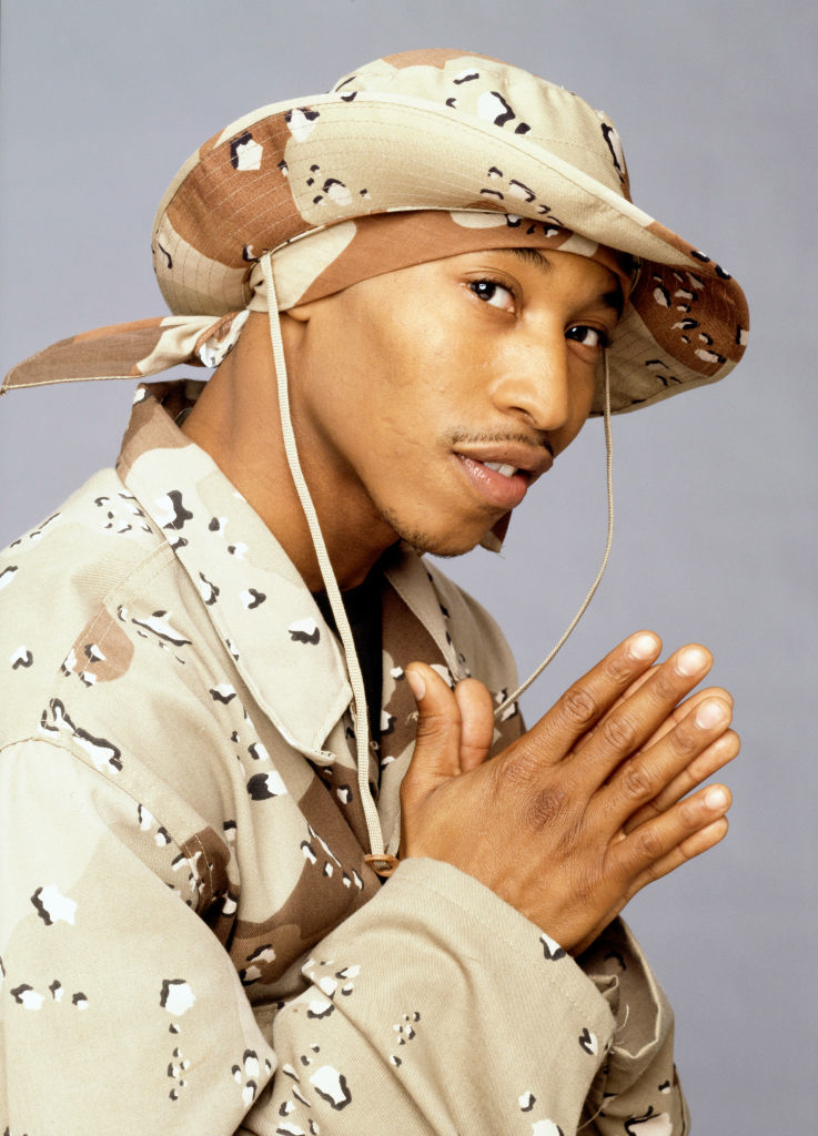 Rakim wearing a desert camouflage jacket and matching hat, posing with hands together in a prayer gesture