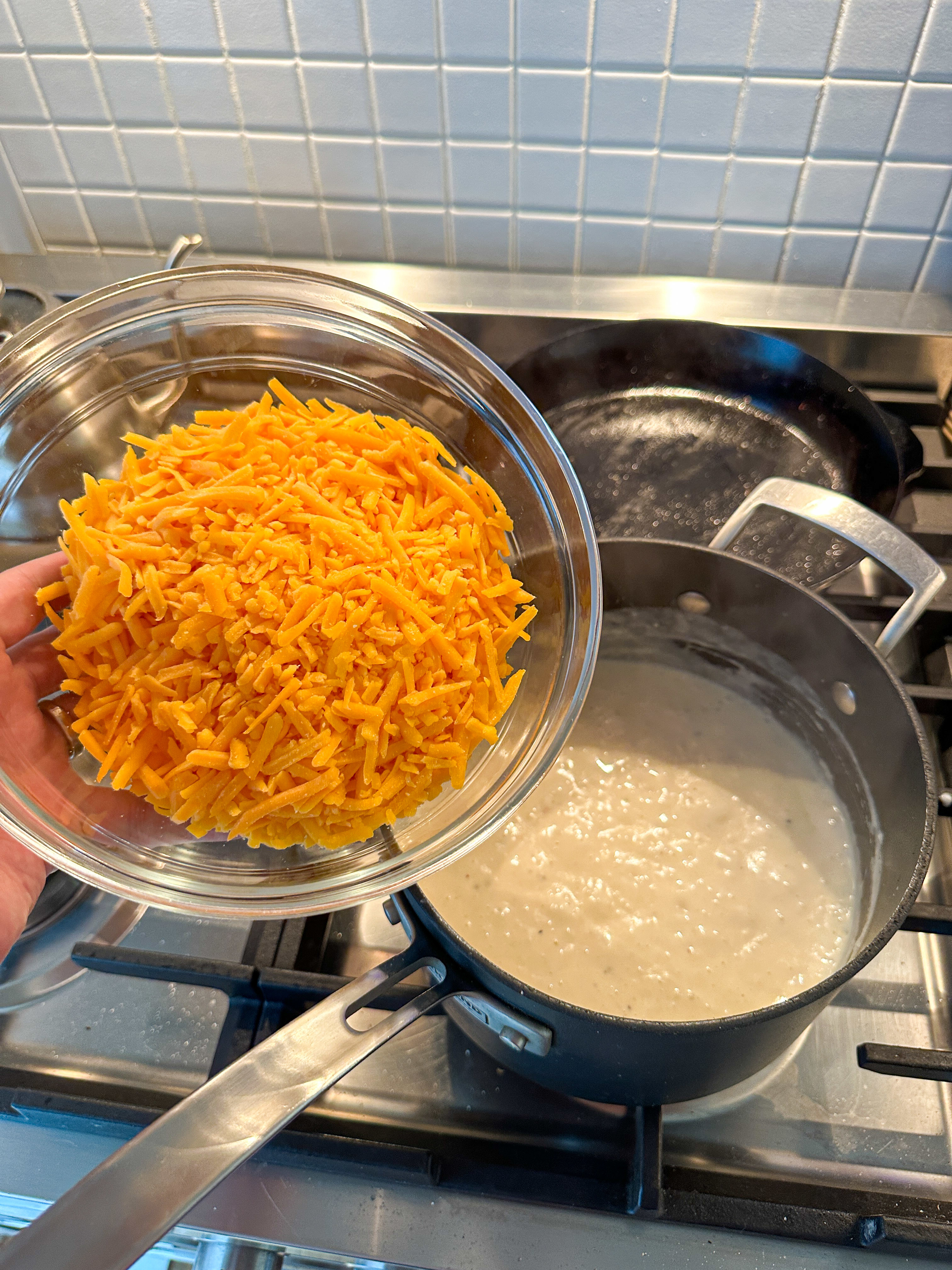 A glass bowl filled with shredded cheese is being held above a pot of creamy sauce on a stovetop, ready to be added into the mixture