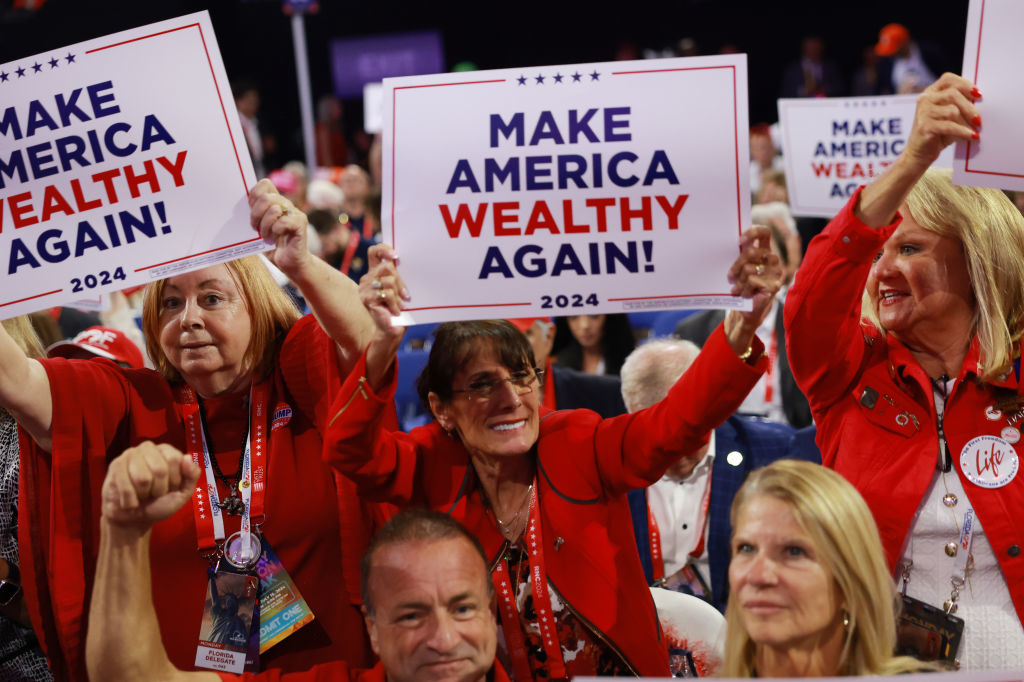 A group of people holds signs reading &quot;Make America Wealthy Again!&quot; and &quot;2024&quot; at a crowded event. They are wearing red attire and appear enthusiastic