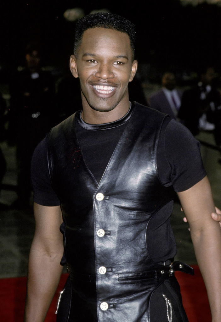 Jamie Foxx smiling and wearing a black leather vest over a black T-shirt at a public event