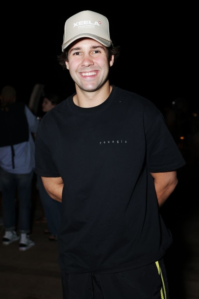David Dobrik smiling, wearing a XCEELA hat and black casual outfit at an event