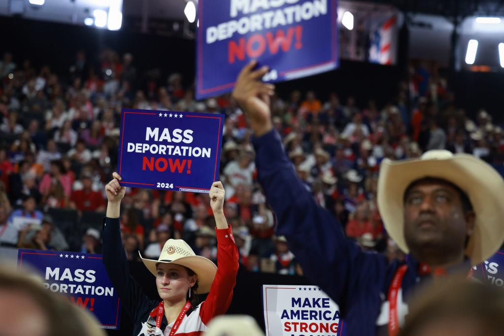 People at a crowded rally hold signs reading &quot;MASS DEPORTATION NOW!&quot; and &quot;MAKE AMERICA STRONG AGAIN.&quot; Several individuals wear cowboy hats