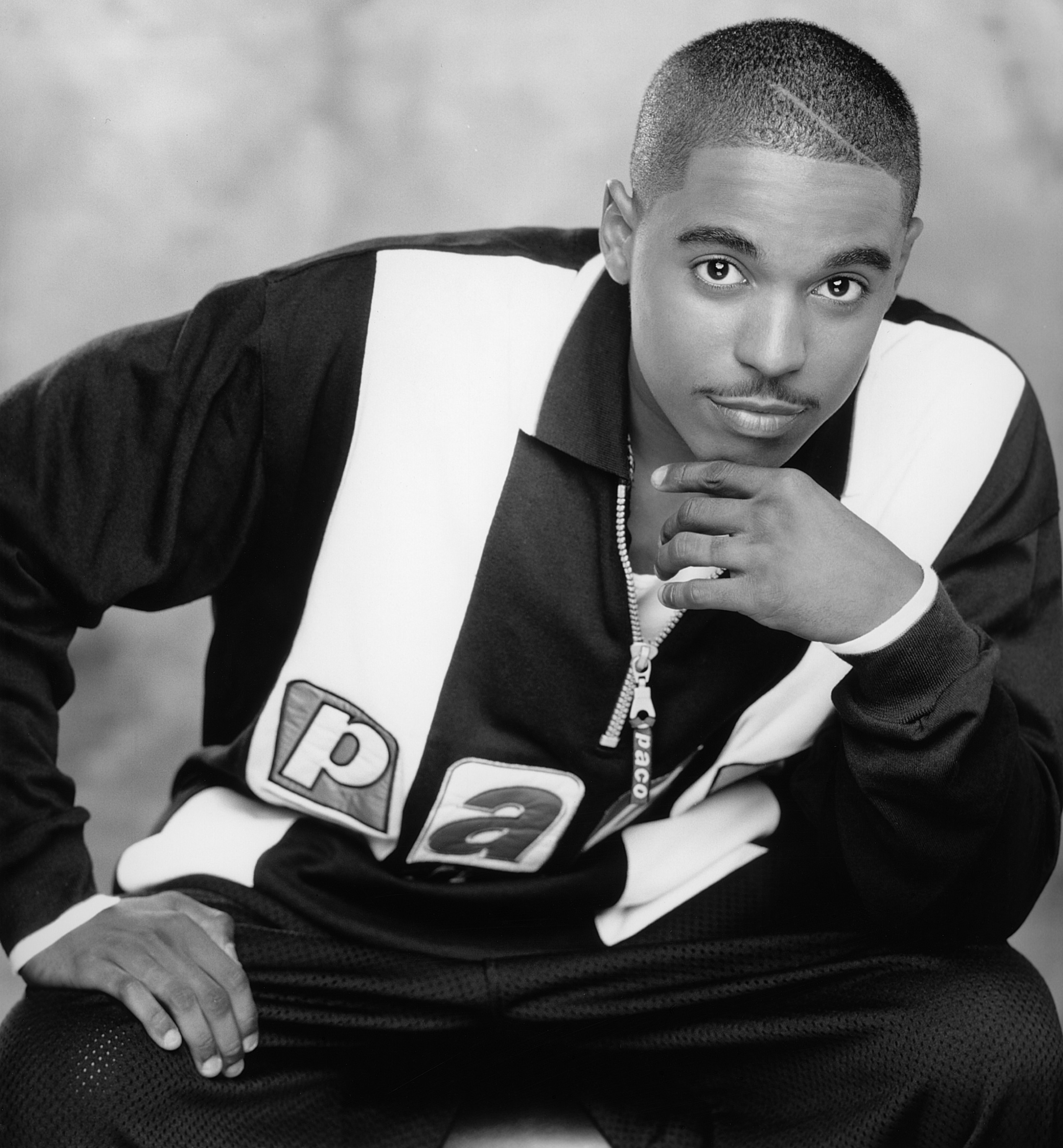 A person is posing with one hand on their chin, wearing a collared shirt featuring a bold graphic &quot;Pa&quot; design