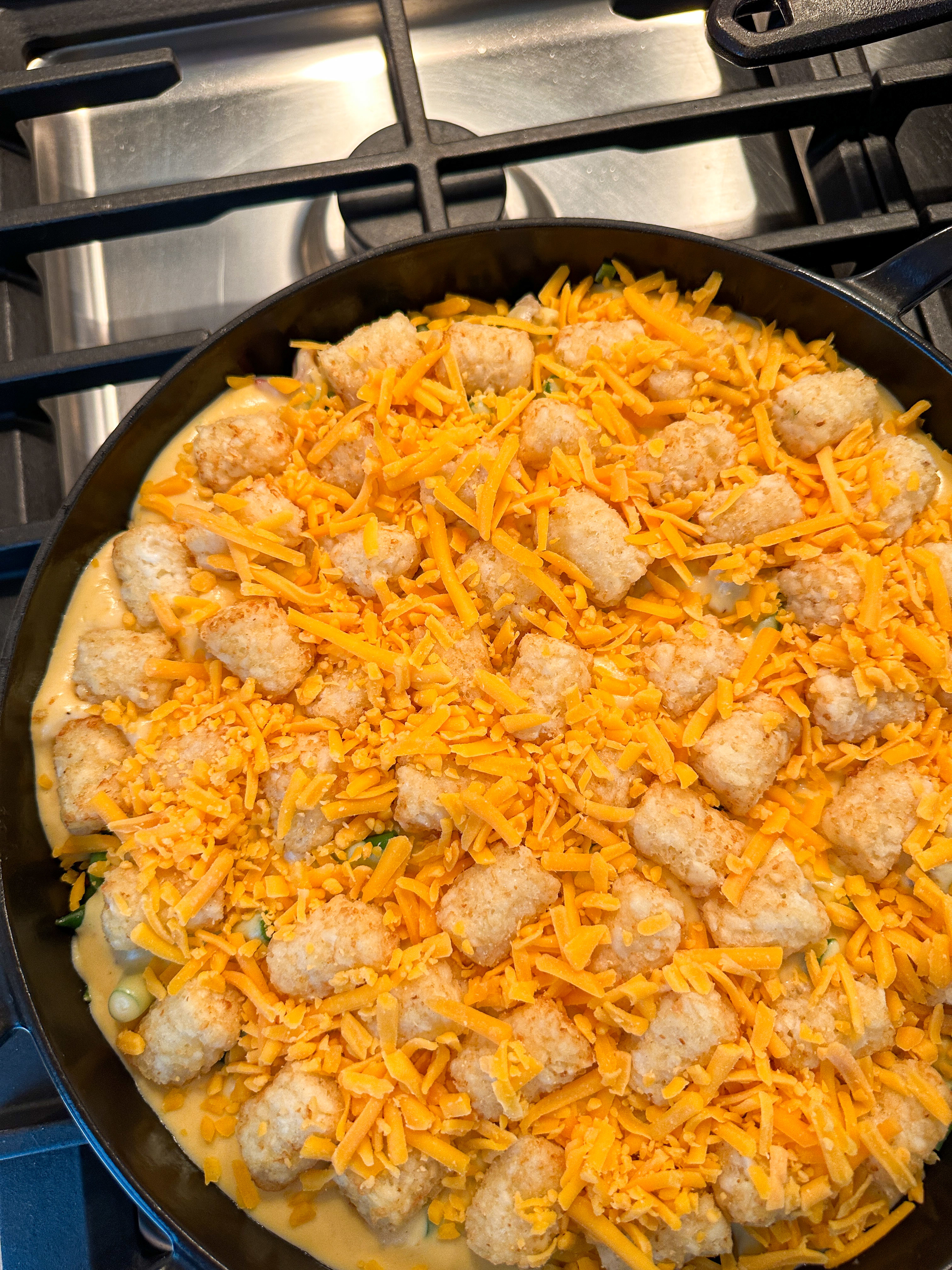 A skillet on a stove filled with a dish of tater tots topped with shredded cheese, ready for baking