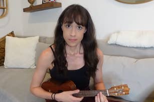 Colleen Ballinger sitting on a couch, playing a ukulele, wearing a black tank top