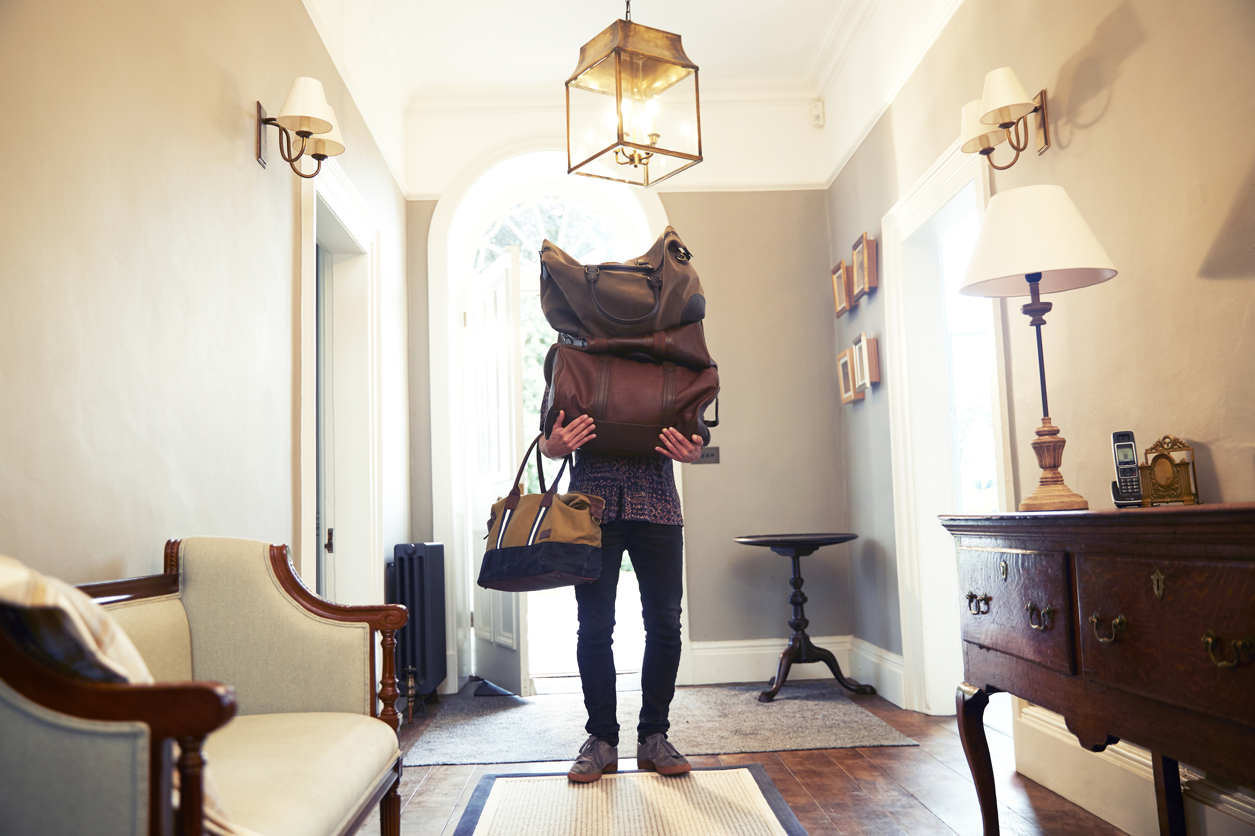 Person in a hallway carrying an exaggeratedly large stack of bags that obscures their face and upper body. Furniture and decor visible in the room