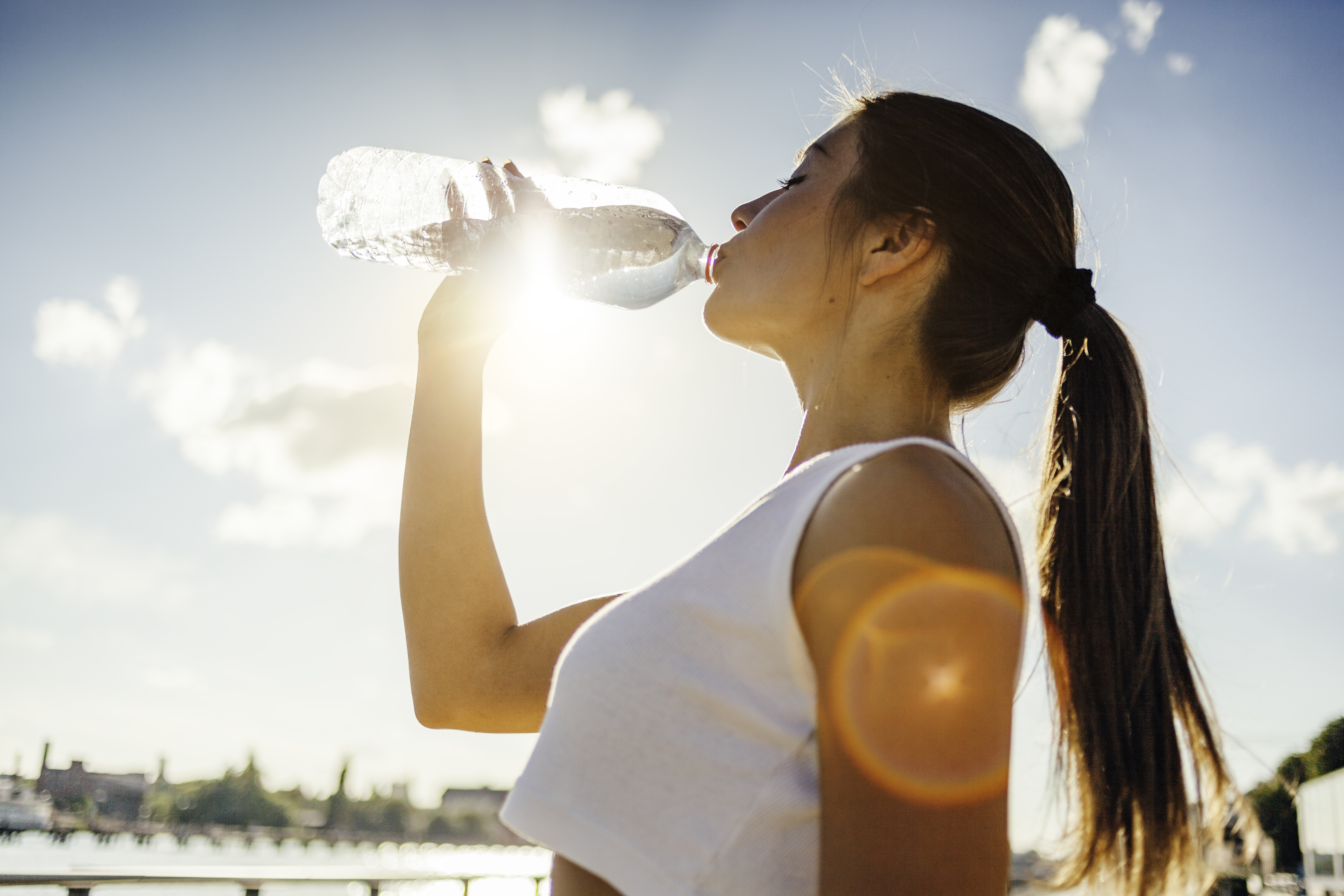A woman with long hair in a ponytail drinks water from a bottle outdoors on a sunny day