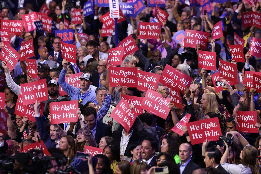 A large crowd holds up red signs that read &quot;We Fight. We Win.&quot; at a rally. People wear casual and business attire