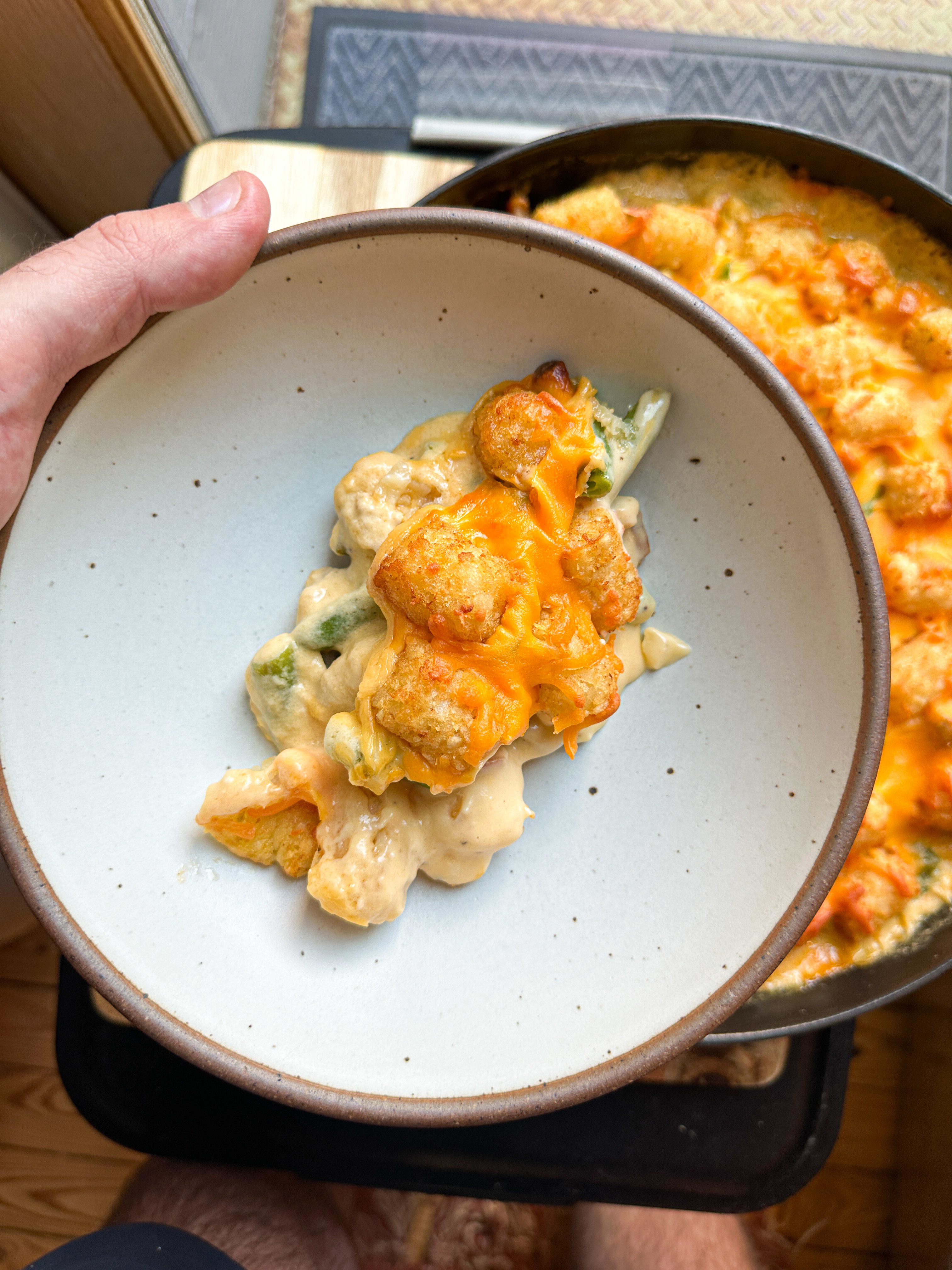 Bowl of cheesy casserole being held above a larger dish of the same casserole