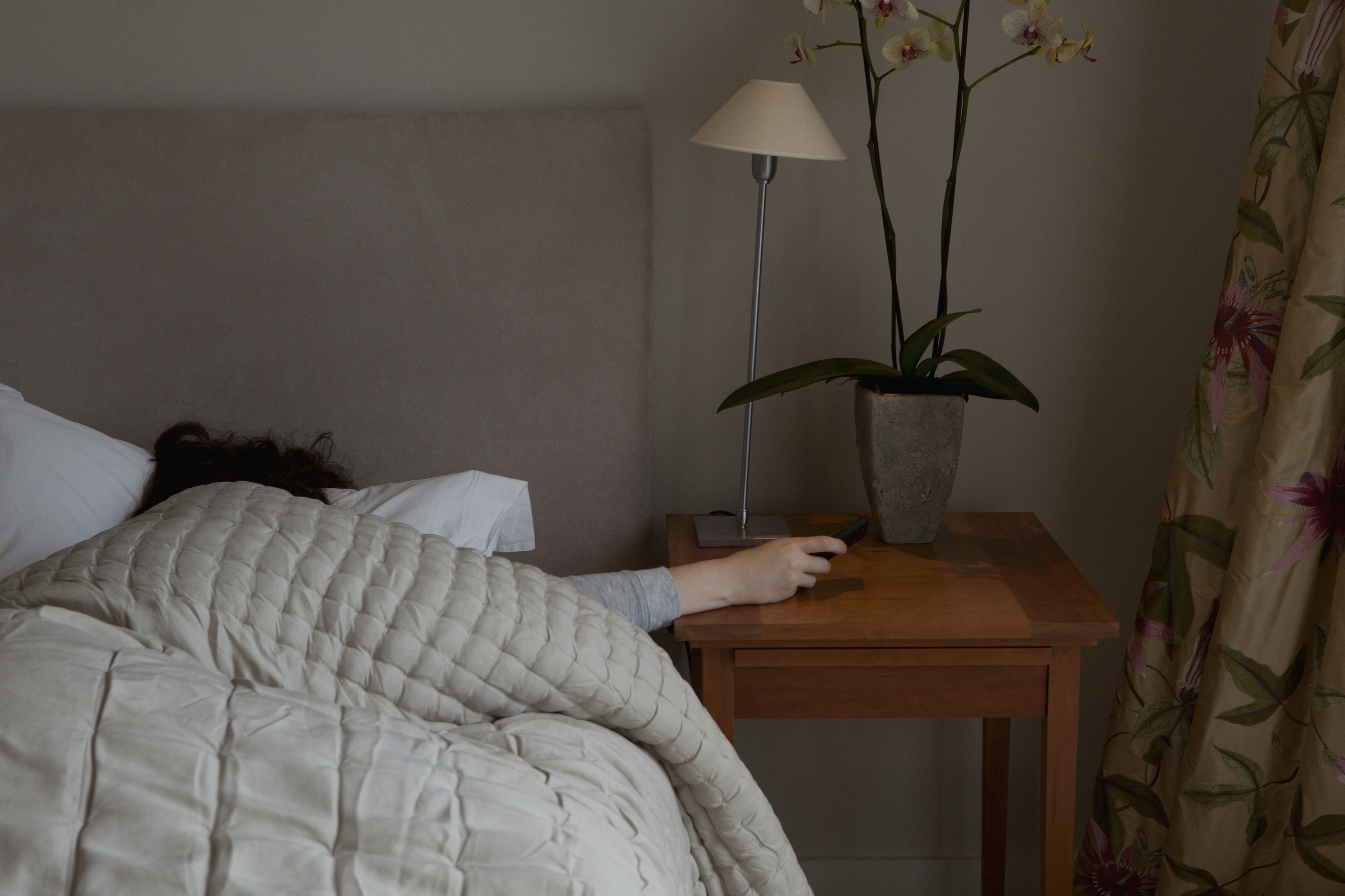 A person lies in bed under a blanket, reaching out to a wooden bedside table with a lamp and a potted orchid