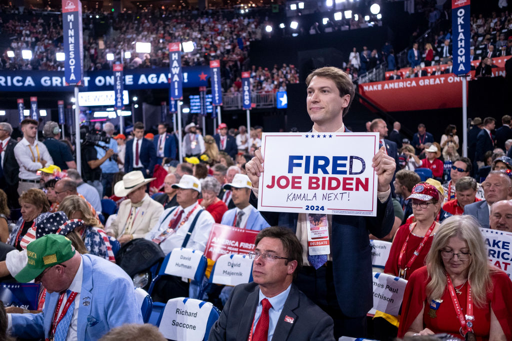 A person holds a sign that reads &quot;Fired Joe Biden, Kamala Next!&quot; at a political rally. The audience is focused on the stage, with people wearing event badges and hats