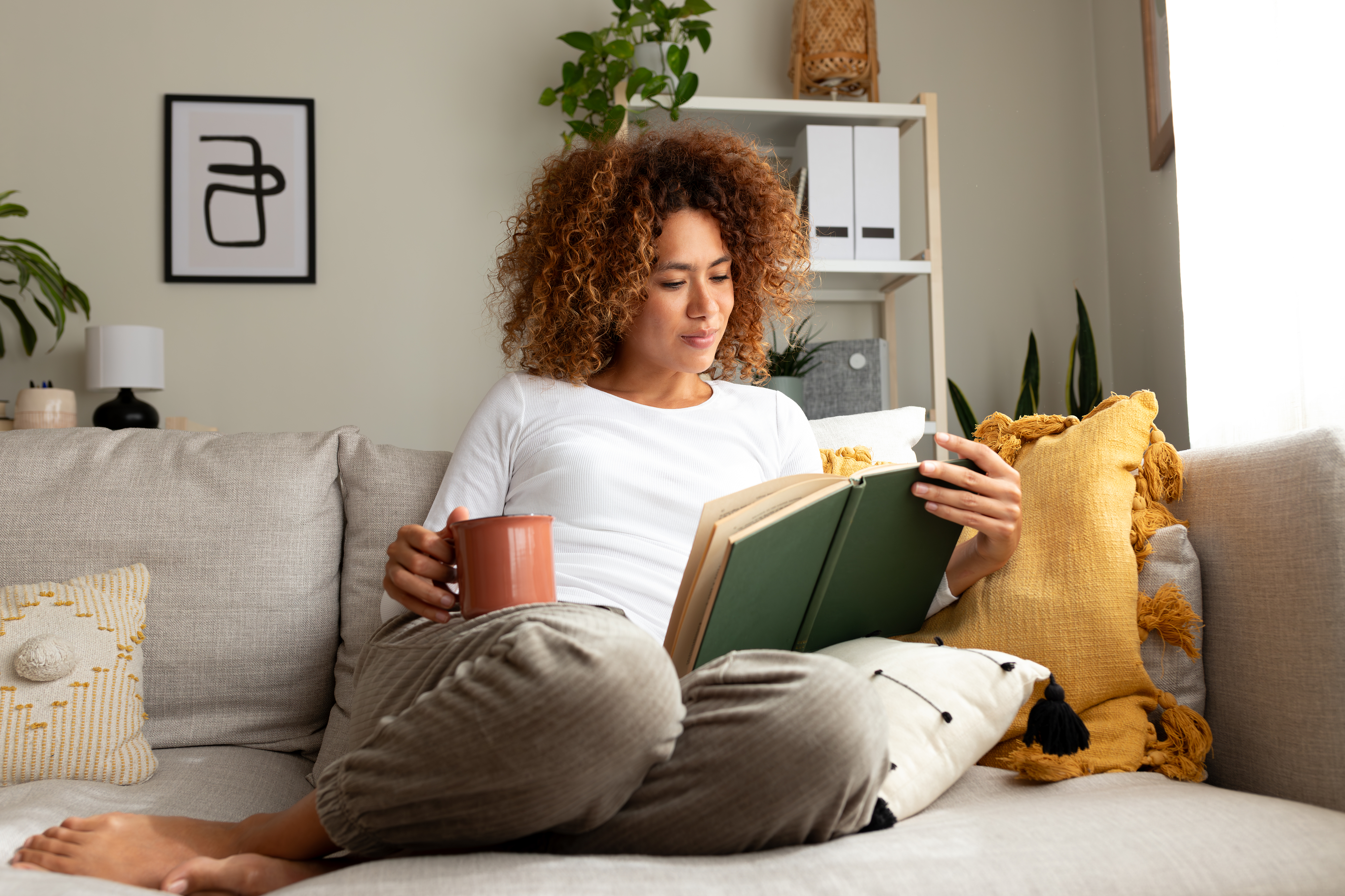 Person sitting on a sofa, reading a book and holding a mug, surrounded by pillows and home decor. They appear relaxed and comfortable