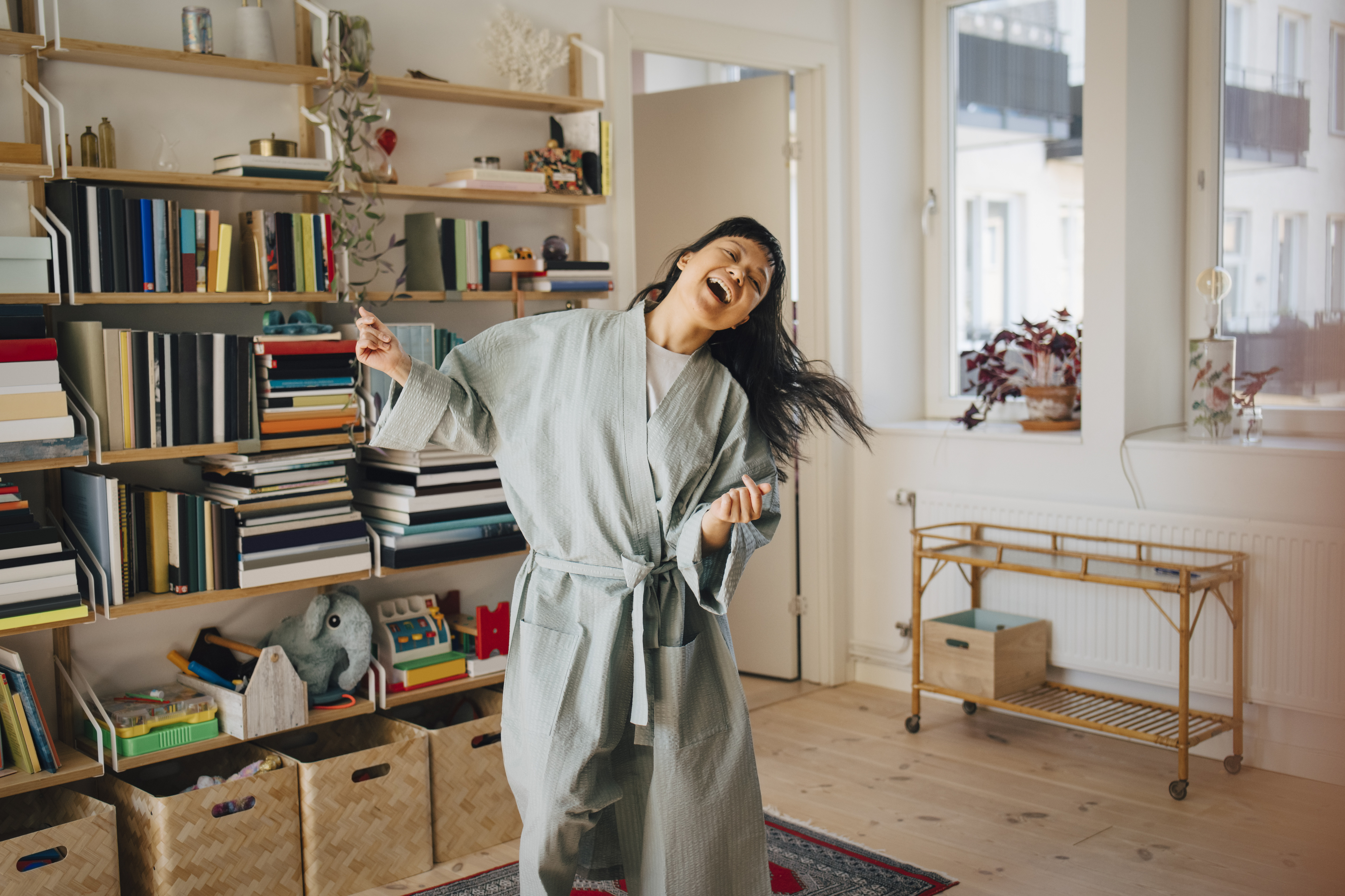 A woman in a comfortable, casual robe joyfully dances in her living room, surrounded by bookshelves, potted plants, and wicker storage baskets