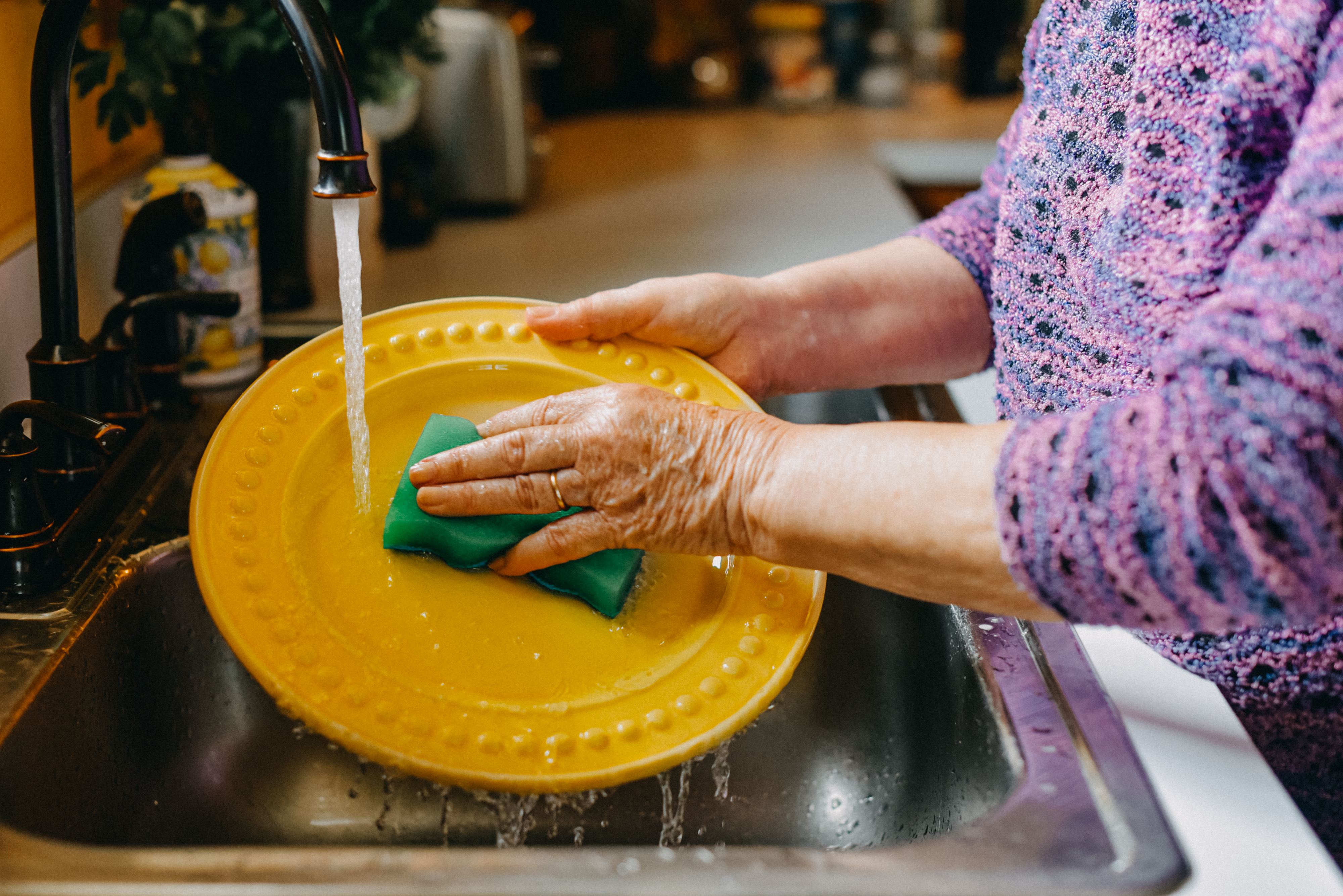 Person washing a yellow plate with a green sponge under running water in a kitchen sink