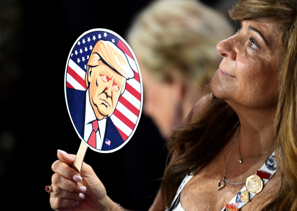 A woman in an audience holds a fan with a cartoon image of Donald Trump against an American flag background. Another person is blurred in the background