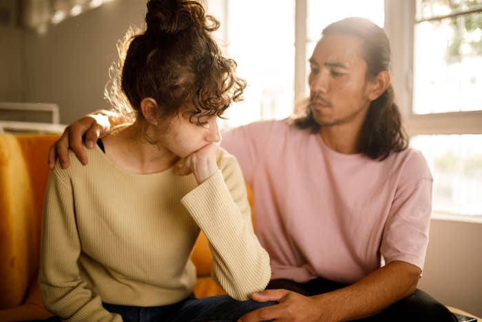 A woman looks sad with her hand to her mouth while a man with long hair sits beside her and puts a comforting hand on her shoulder