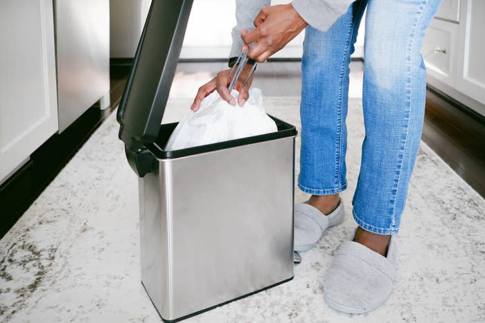 Person in jeans and slippers putting a trash bag into a stainless steel kitchen trash can