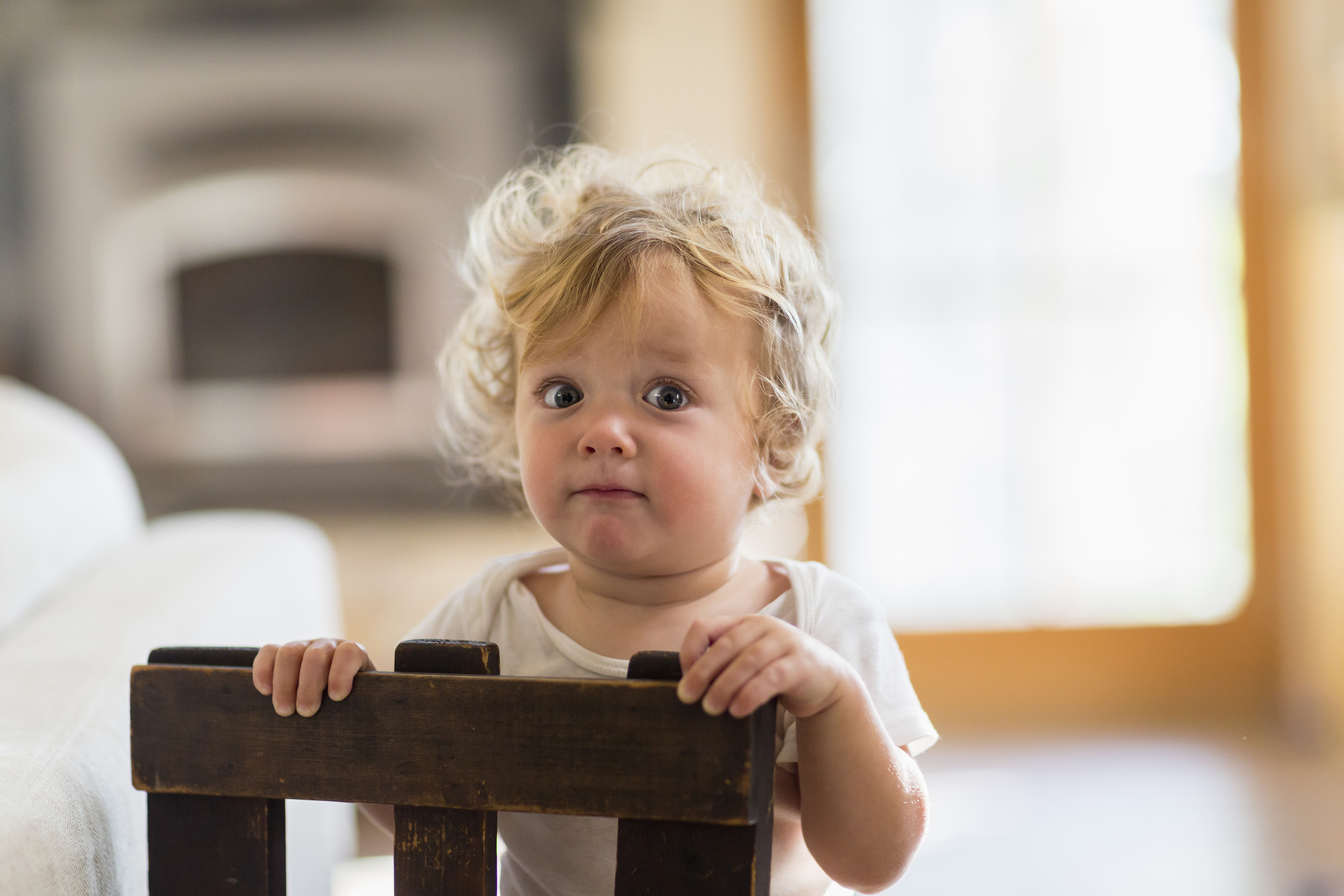 A young child with curly hair stands behind a dark wooden chair, looking up with a curious expression in a warmly lit room