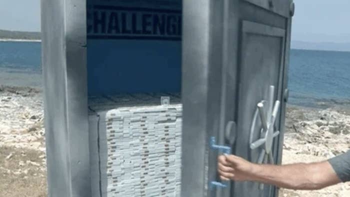 A person is opening the door to a large safe with stacks of money inside, near a scenic outdoor location by the water
