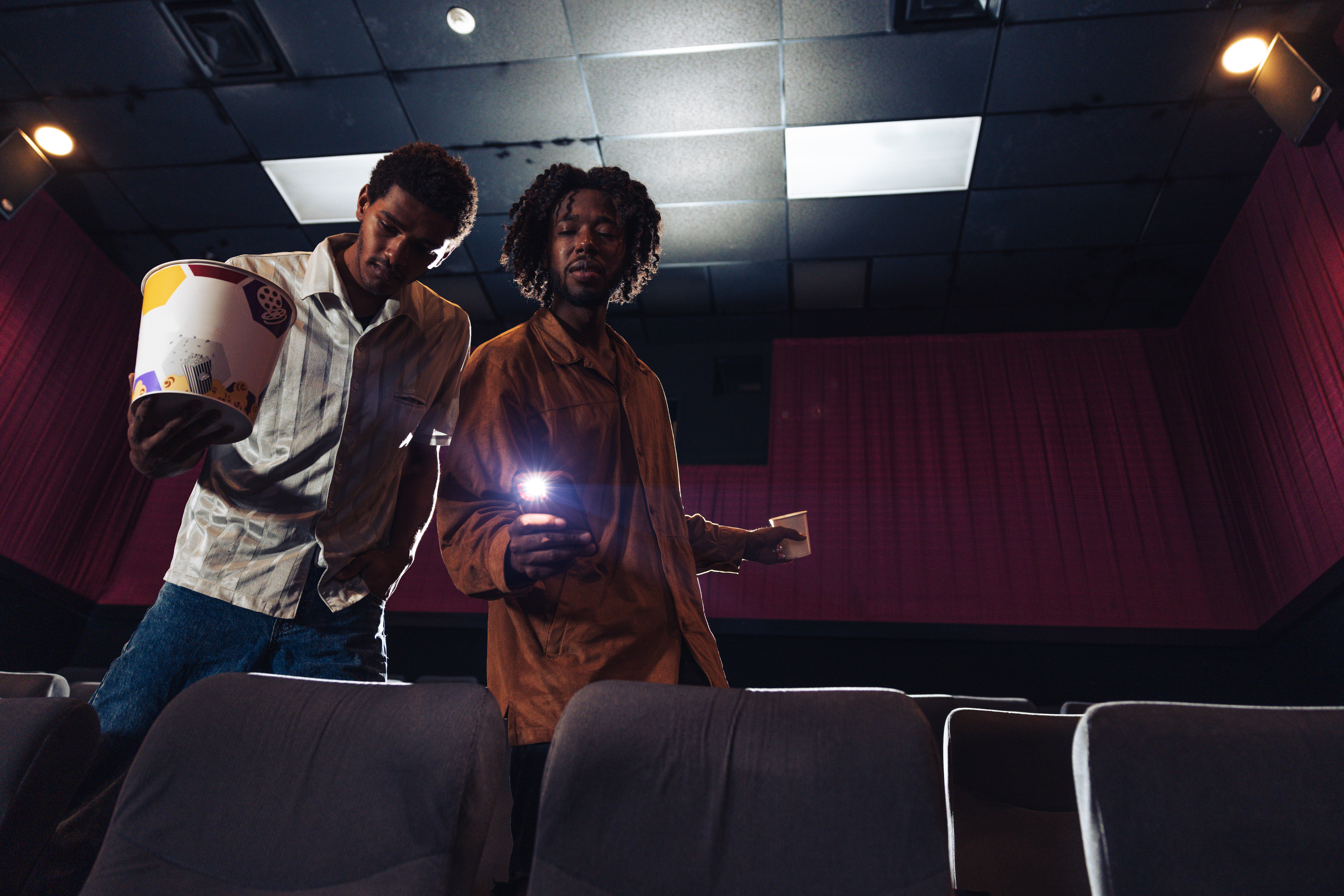 Two men, one holding a bucket of popcorn and the other shining a flashlight, stand in an empty movie theater