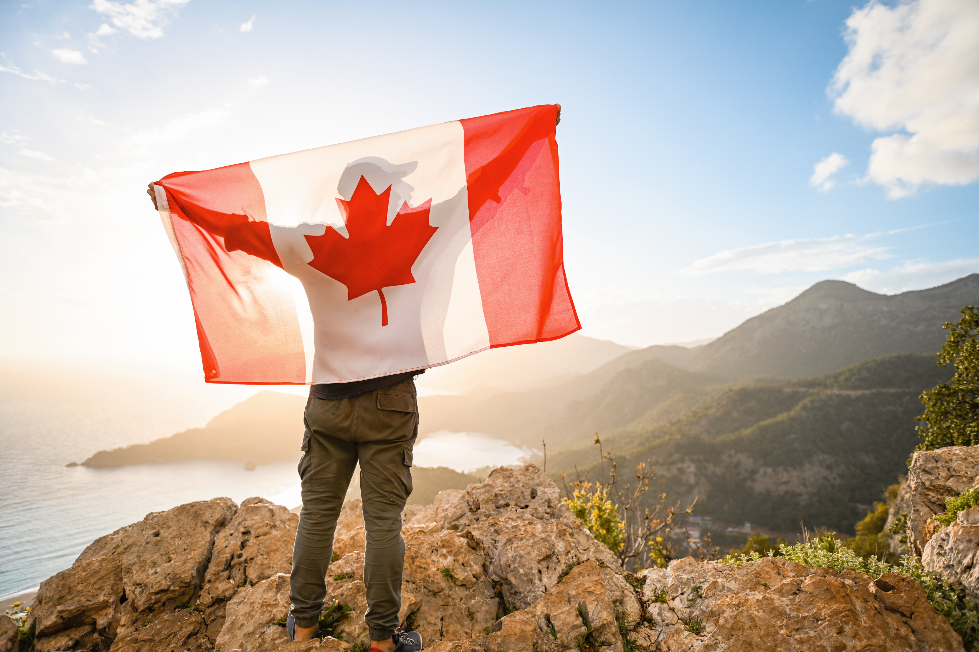 Person holding a large Canadian flag while standing on a rocky cliff, facing a scenic, mountainous landscape