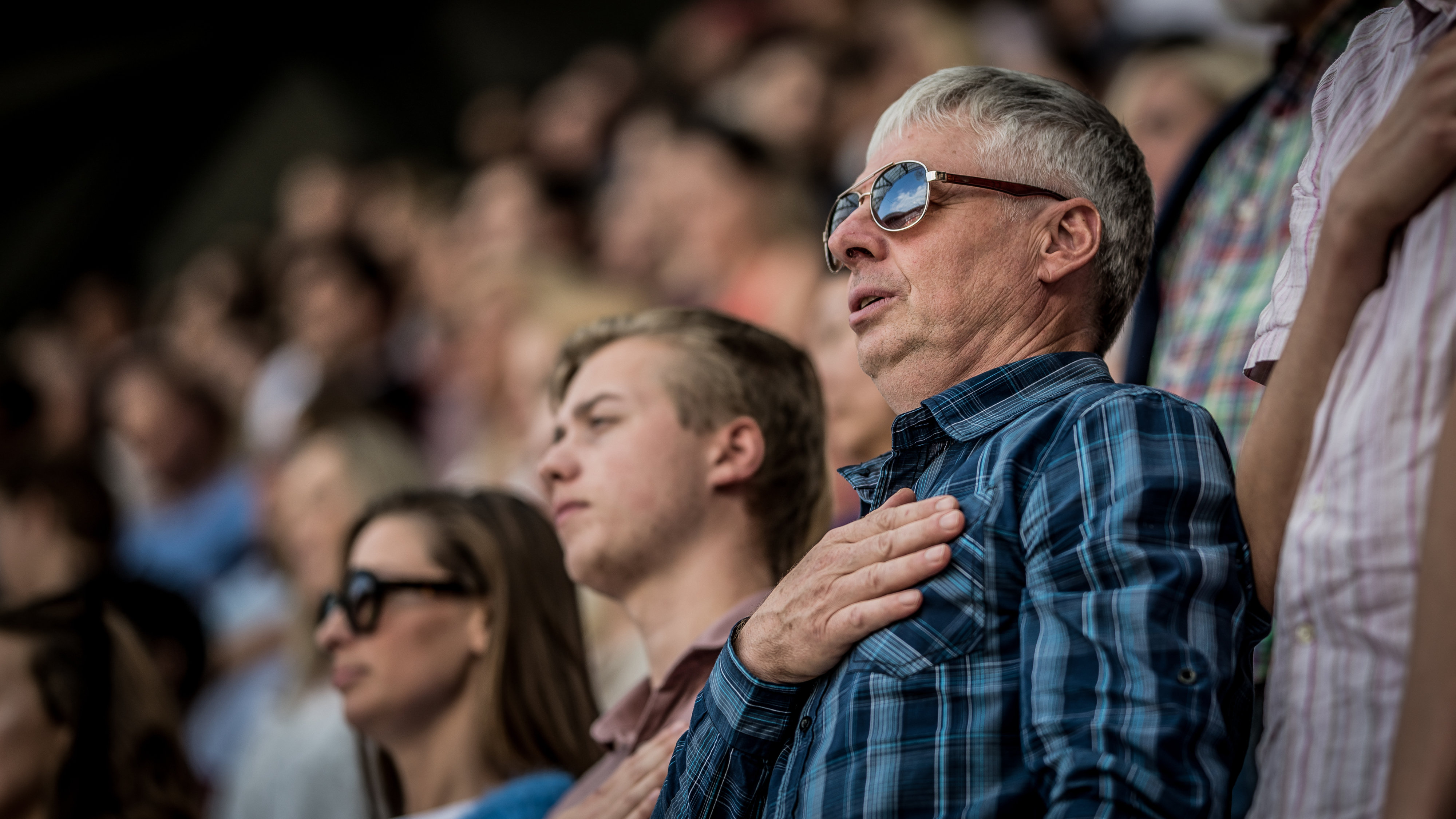 A group of people, including a man in a plaid shirt with his hand over his heart, stands in a crowded stadium