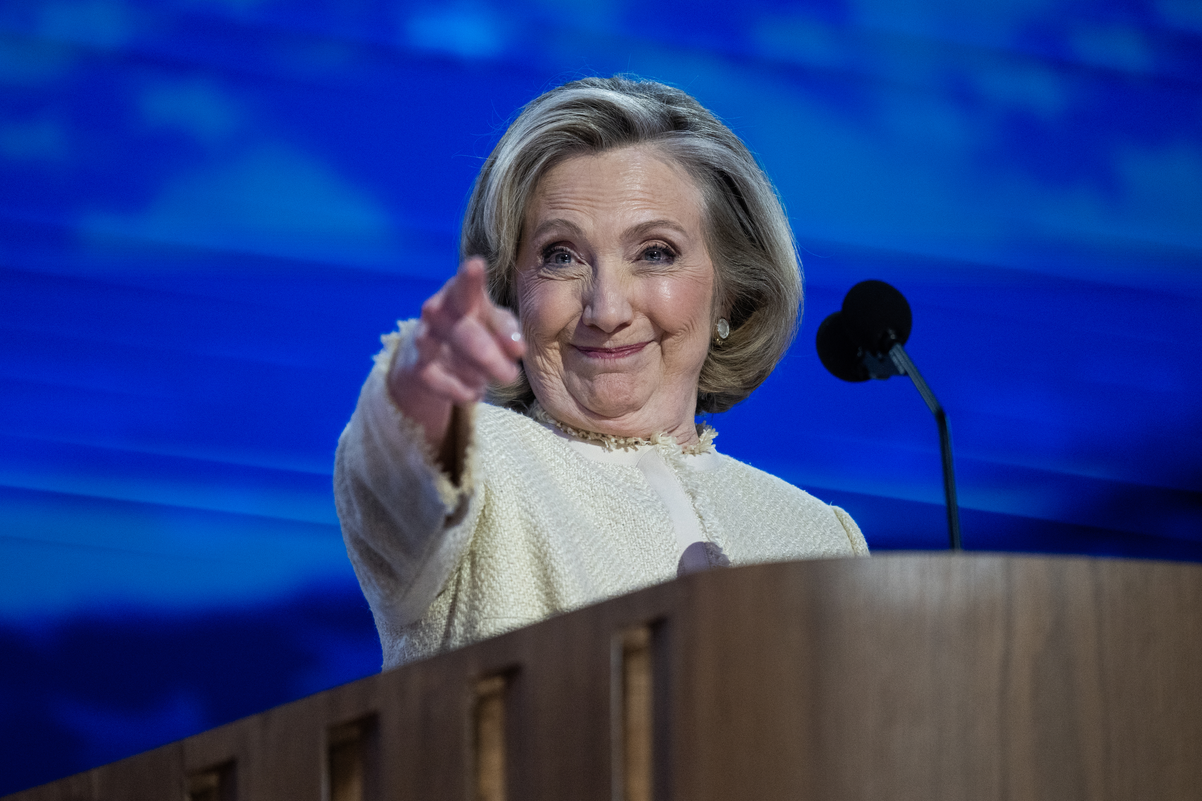 Hillary Clinton, wearing a light jacket, stands at a podium with a microphone, pointing and smiling during a speech